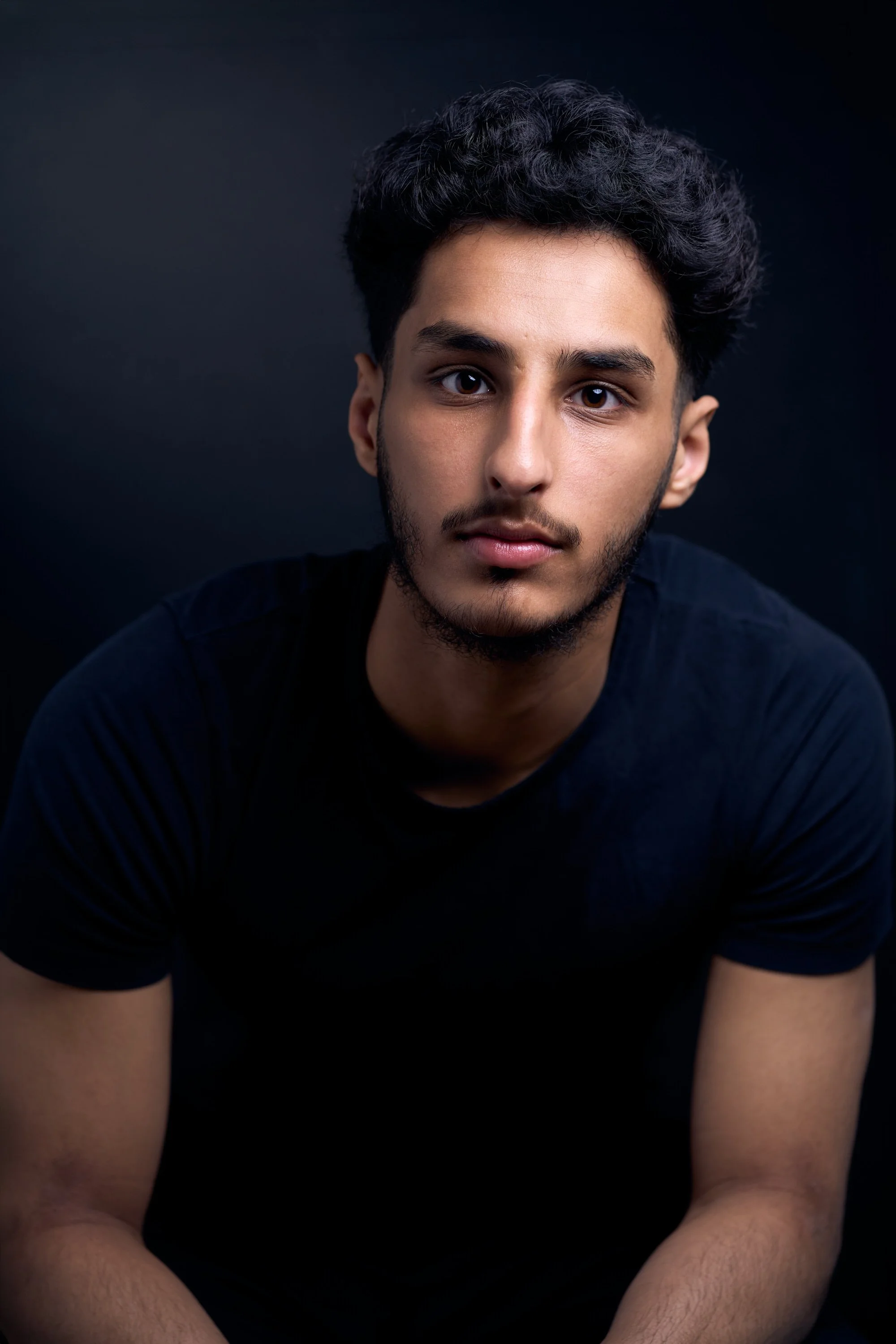 A young man with dark, curly hair and a beard looking at the camera, wearing a black shirt against a dark background.
