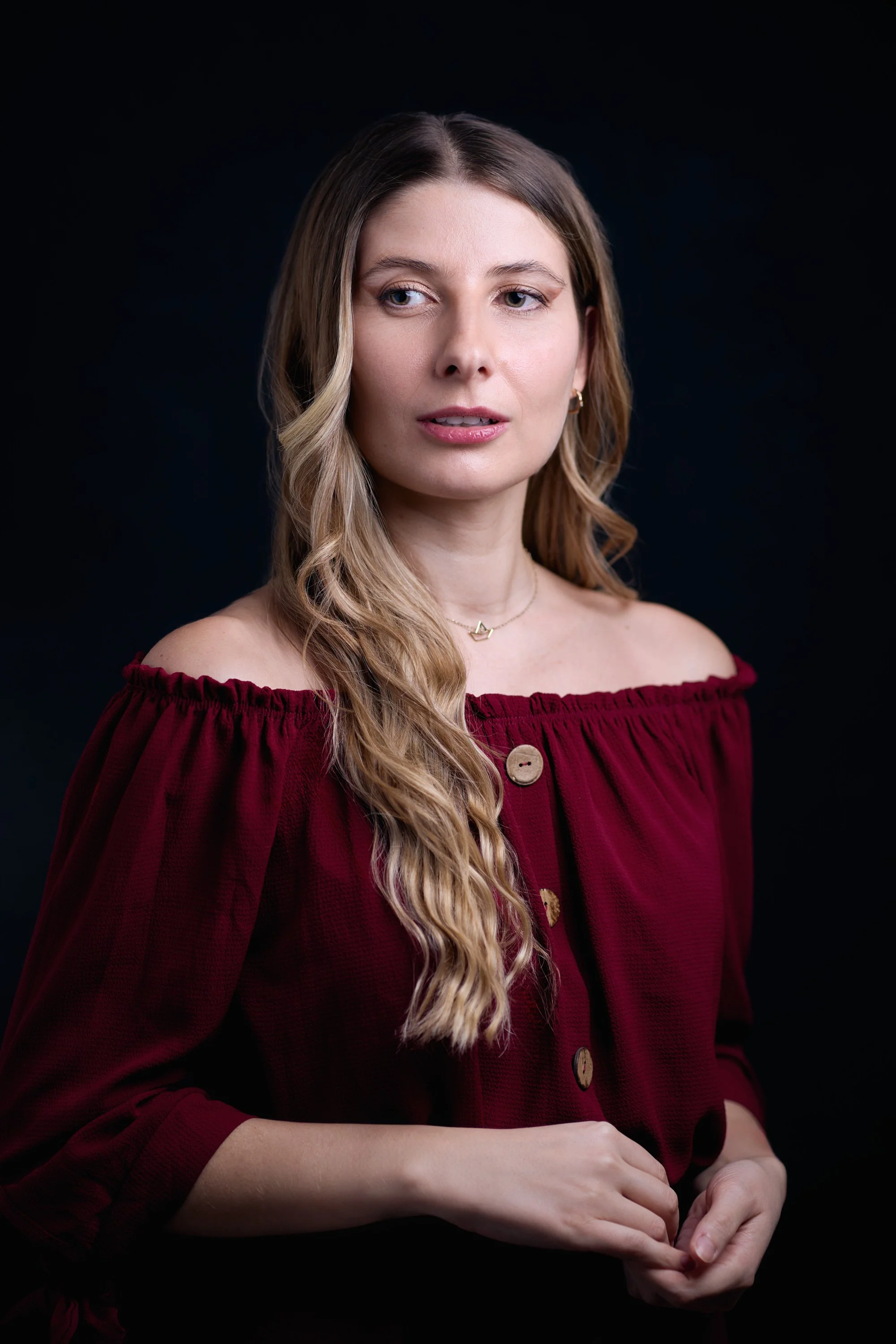 A woman with long, wavy blonde hair wearing a maroon off-shoulder top with buttons and a gold necklace, looking to her right against a black background.