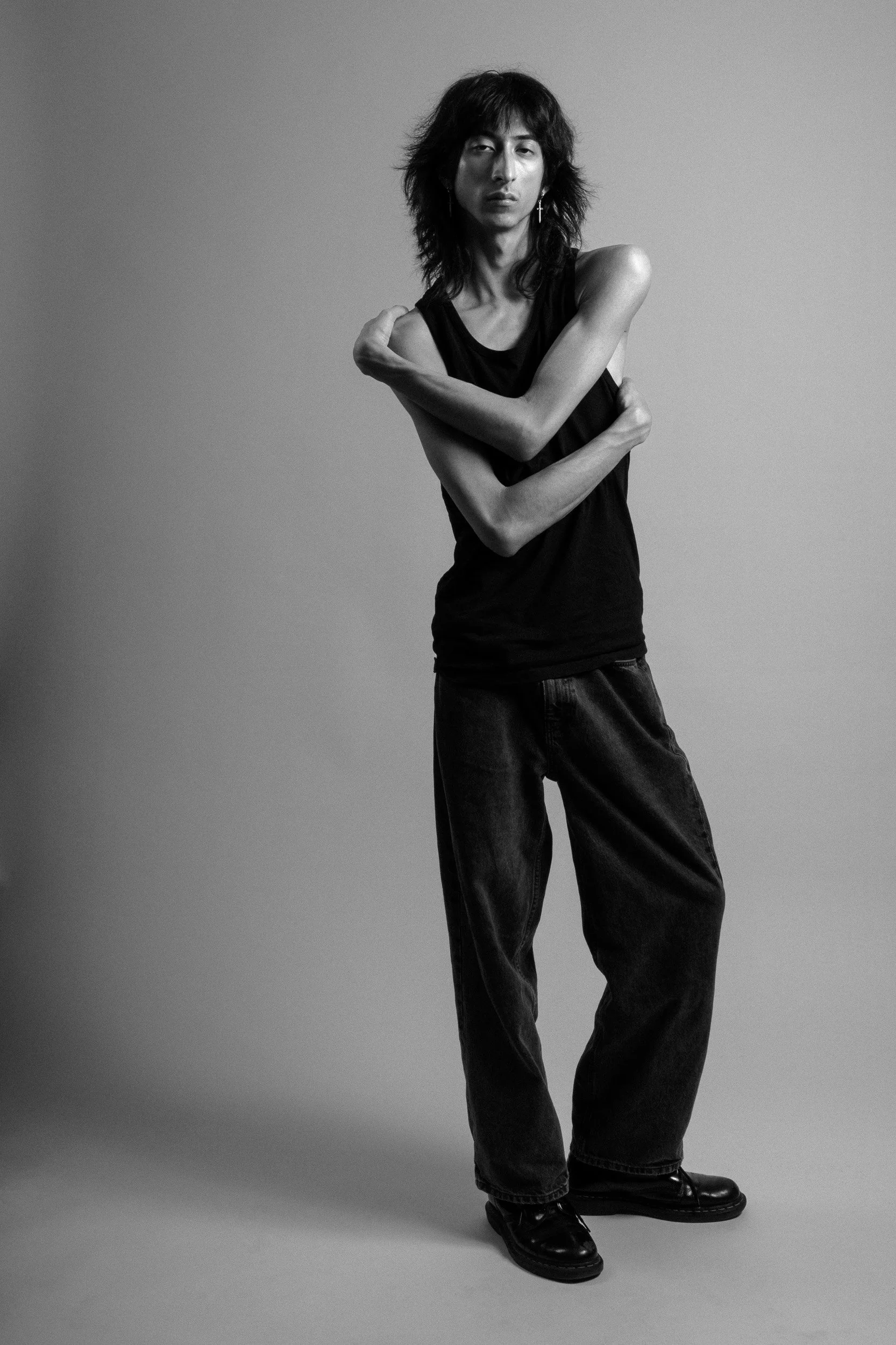 A black and white photo of a woman with shoulder-length hair wearing a sleeveless black top, loose jeans, and shoes, standing against a plain background with arms crossed in front of her chest.
