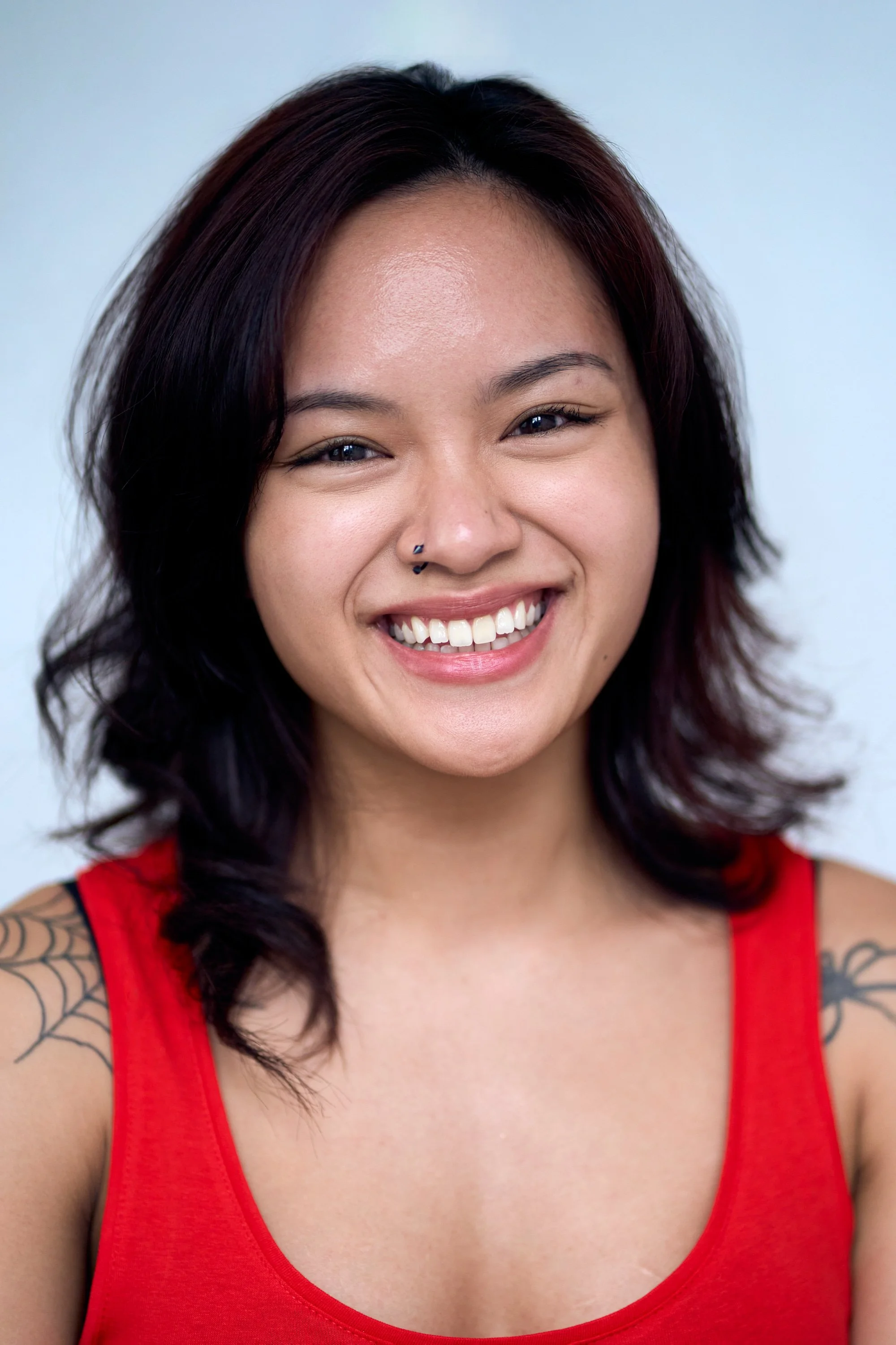 A young woman with dark and reddish hair, smiling broadly, wearing a red tank top, showing part of a tattoo on her right shoulder, against a plain light background.