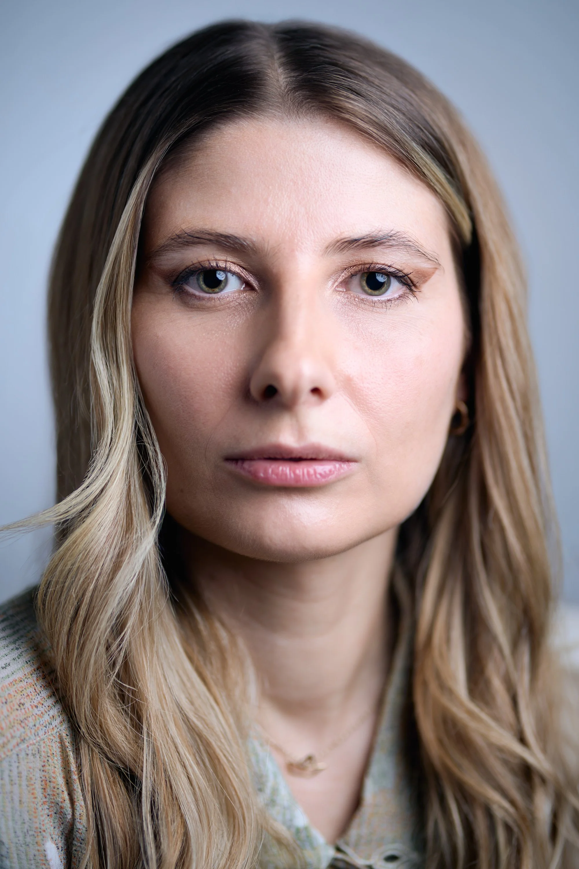 Close-up of a woman with long, wavy blonde hair and bright hazel eyes, wearing a pastel-colored shirt with a subtle pattern, against a plain light background.