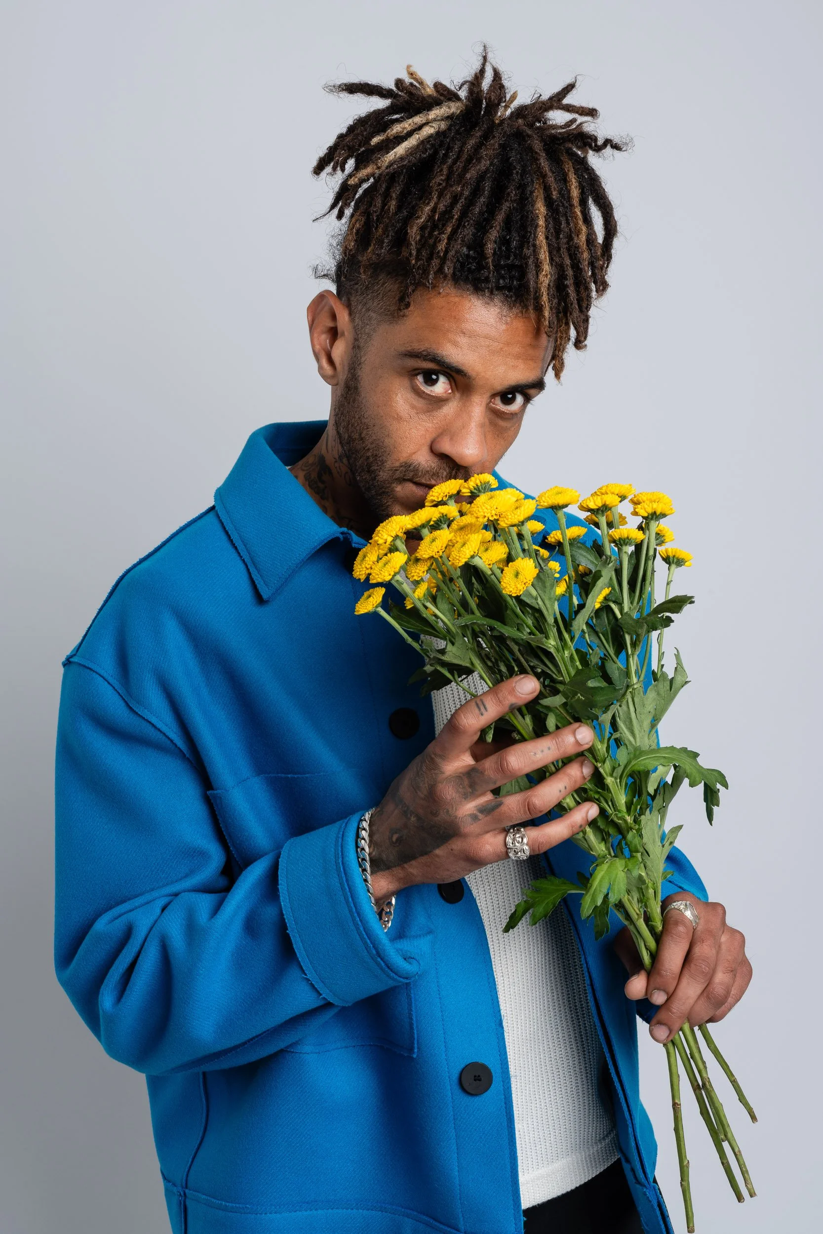 A man with dreadlocks wearing a blue jacket holding a bouquet of yellow flowers against a plain gray background.