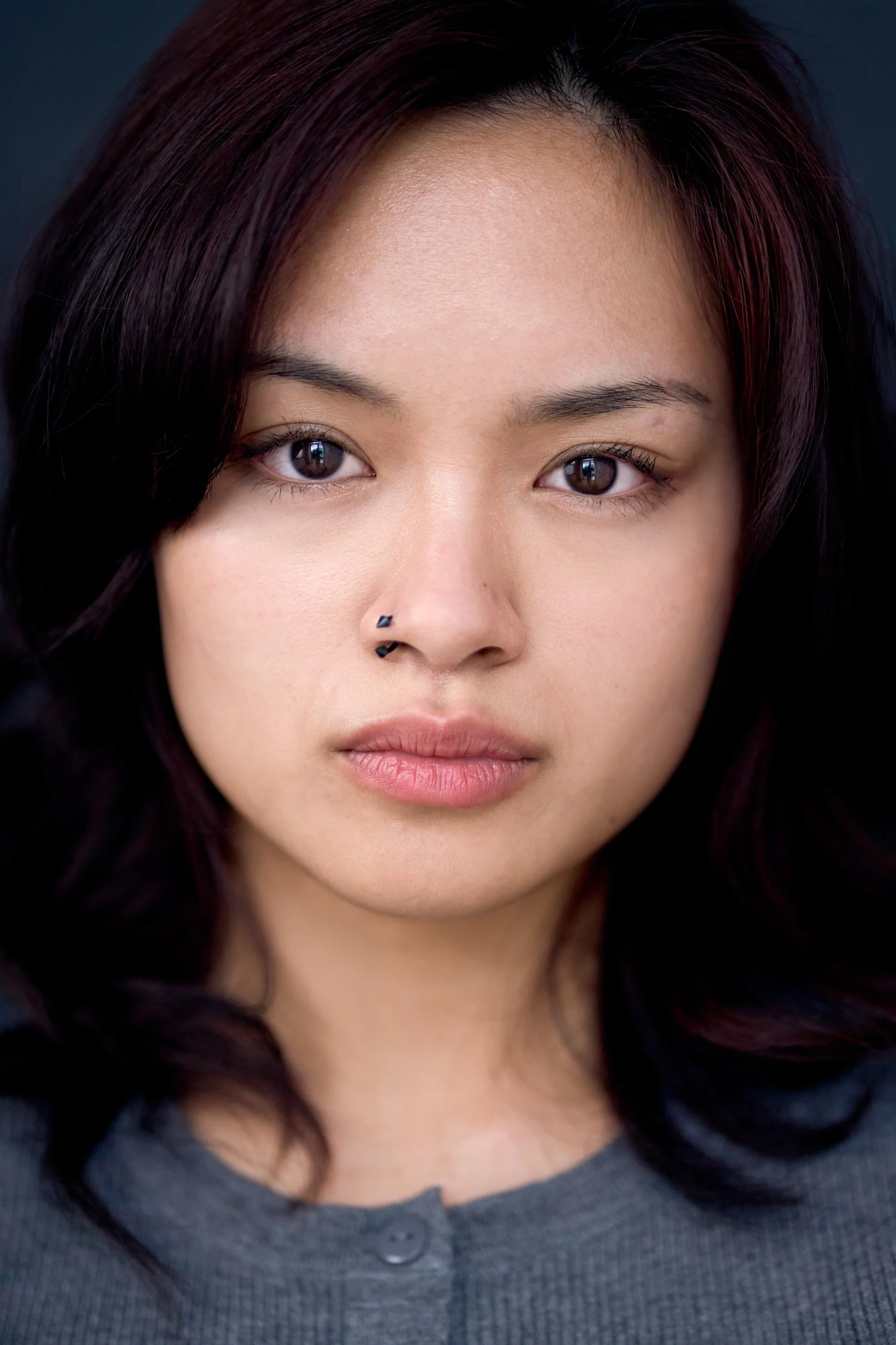 Close-up of a woman's face with dark hair, brown eyes, and a nose piercing, wearing a gray shirt.
