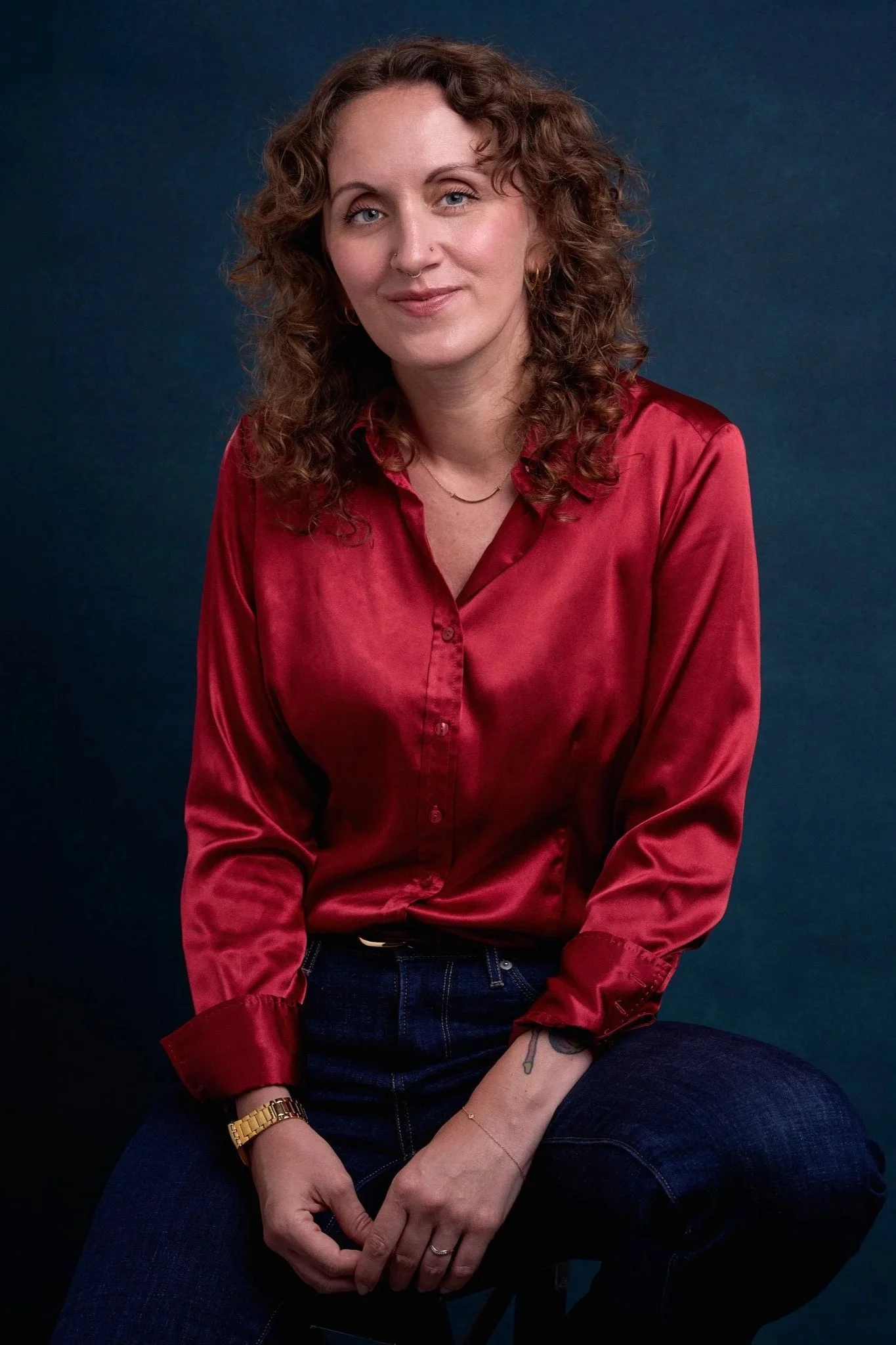 A woman with curly hair, wearing a red silk shirt, dark jeans, a gold wristwatch, and multiple jewelry items, sitting against a dark blue background.