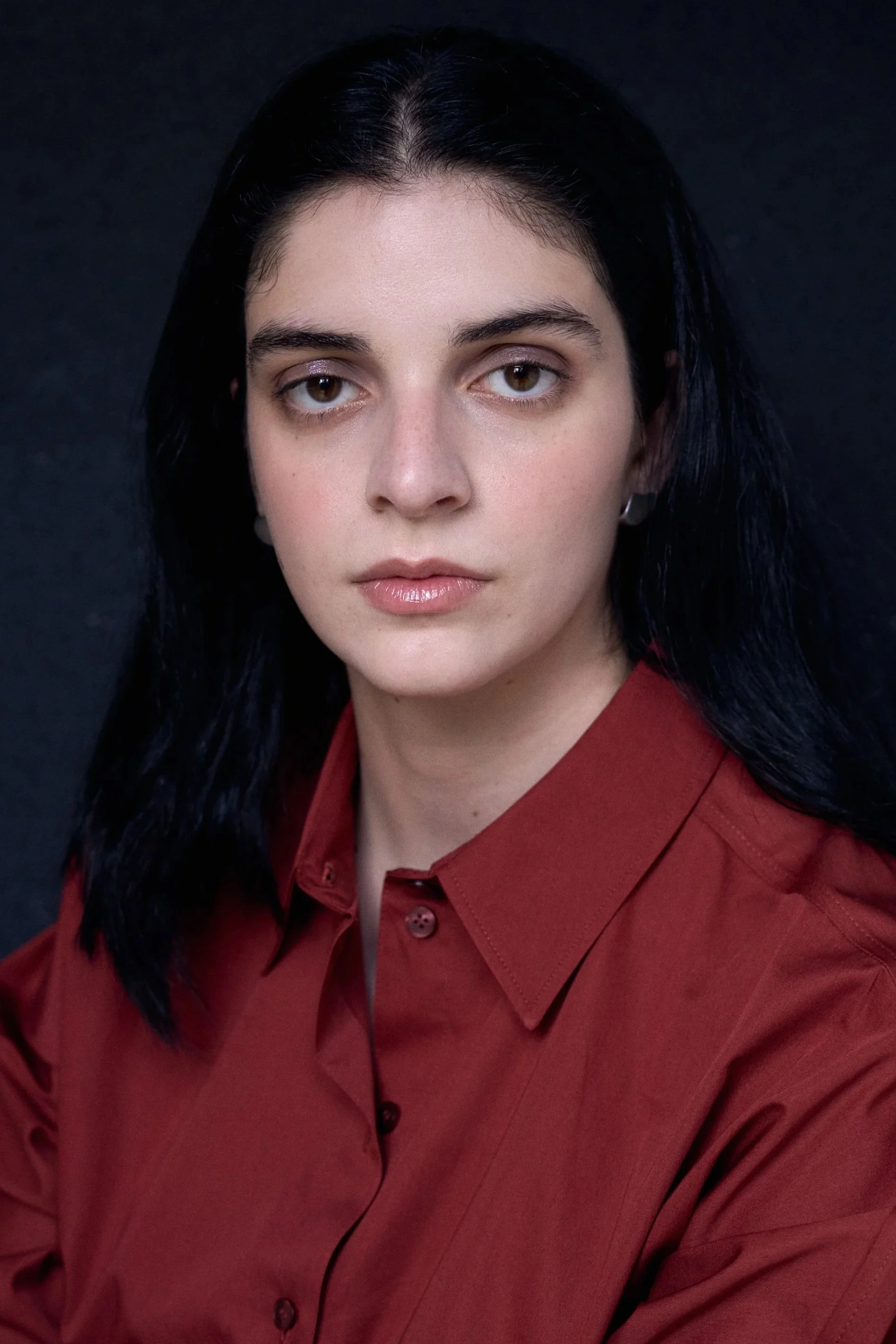 A woman with black hair, wearing a red shirt, looking directly at the camera against a dark background.