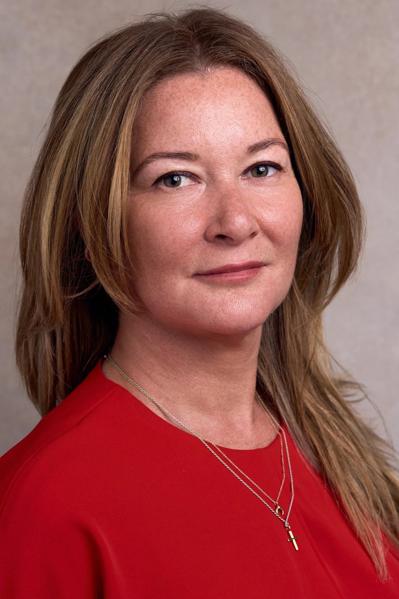 Close-up photo of a woman with long light brown hair, wearing a red top and a silver necklace with a cross pendant. She has light skin, blue eyes, and a neutral expression.