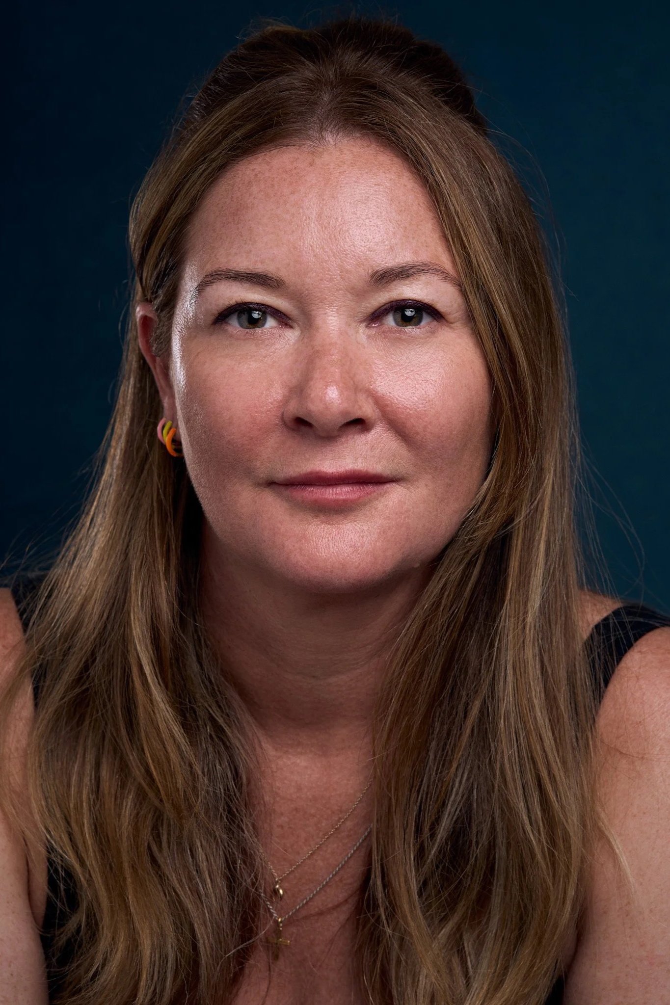 Close-up of a woman's face with long, light brown hair, blue eyes, and wearing colorful earrings.