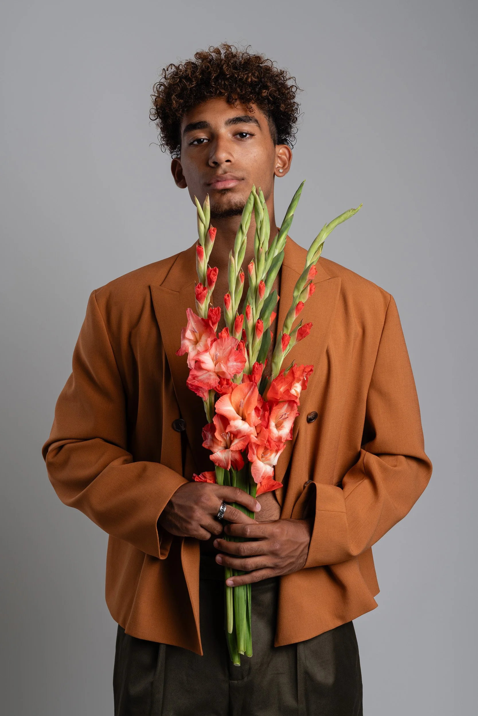 A young man with curly hair dressed in a brown blazer holding a bouquet of red and orange gladiolus flowers against a plain gray background.