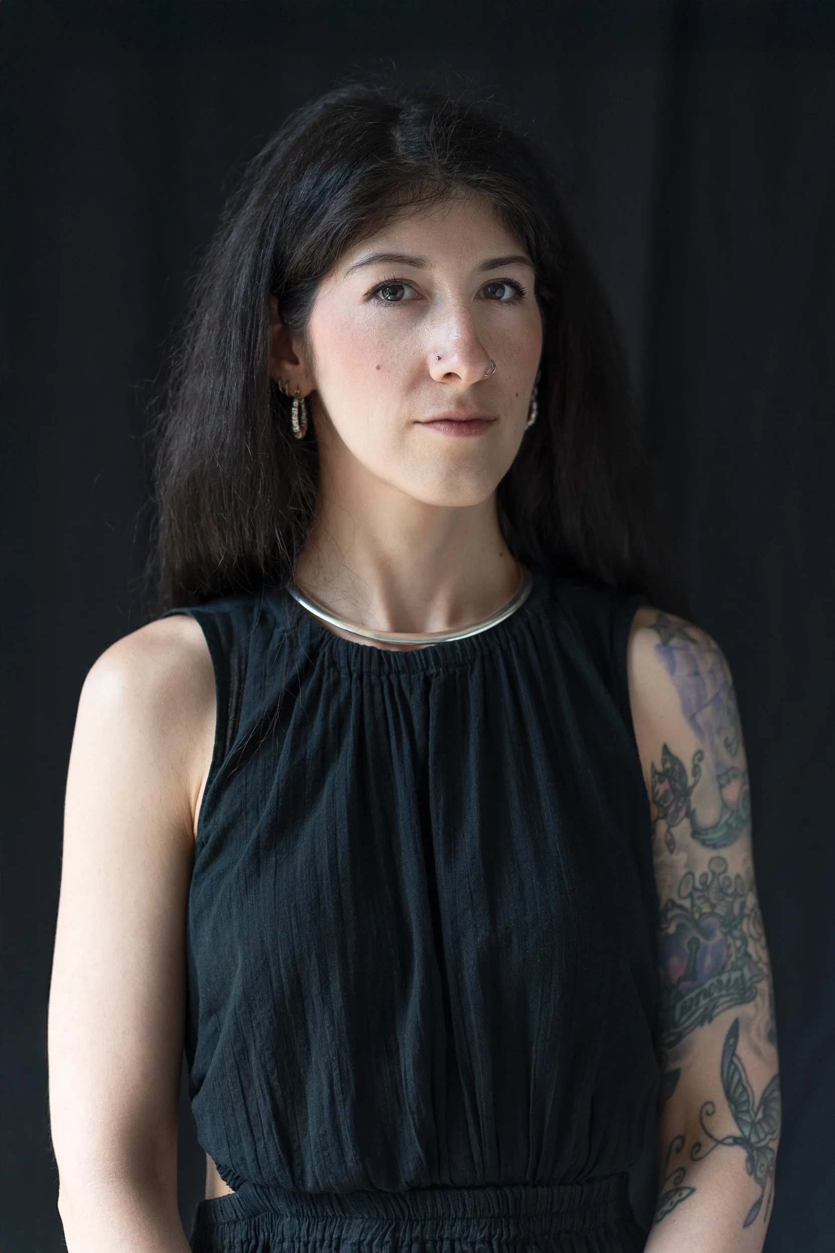 A woman with dark hair and tattoos wearing a black sleeveless top and silver jewelry against a dark background.
