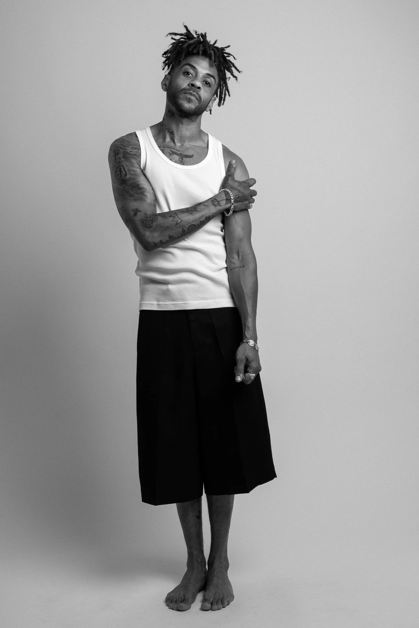 A man with dreadlocks, tattoos, a white tank top, and knee-length shorts standing barefoot against a plain background.