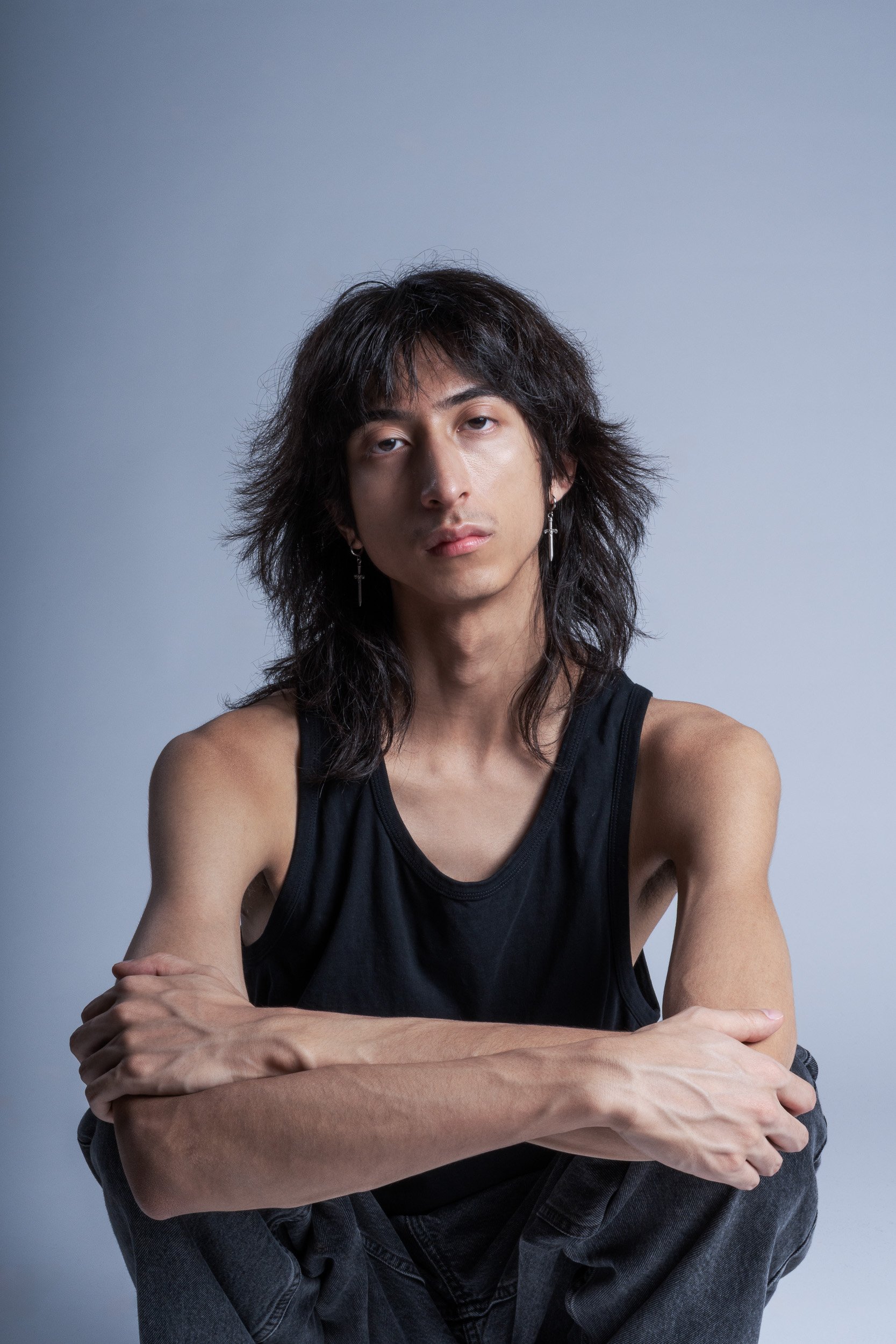 A young man with long, dark hair and earrings, wearing a black tank top, sitting with arms crossed against a plain gray background.