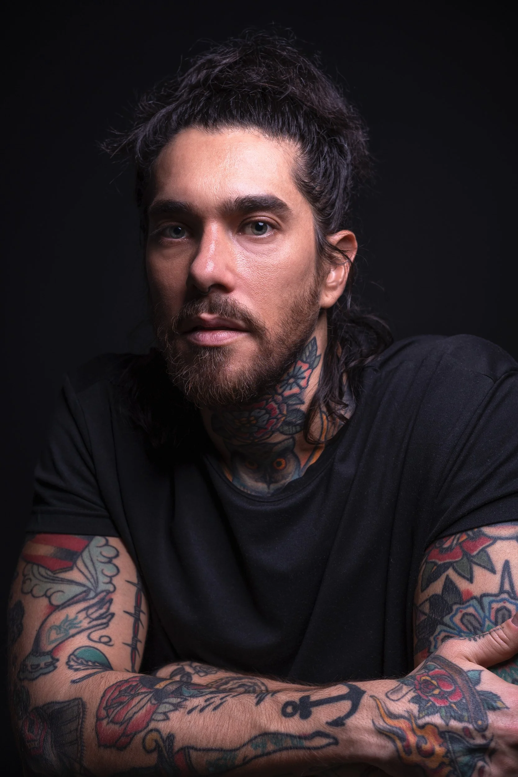 Portrait of a tattooed man with long dark hair tied back, wearing a black shirt, against a dark background.