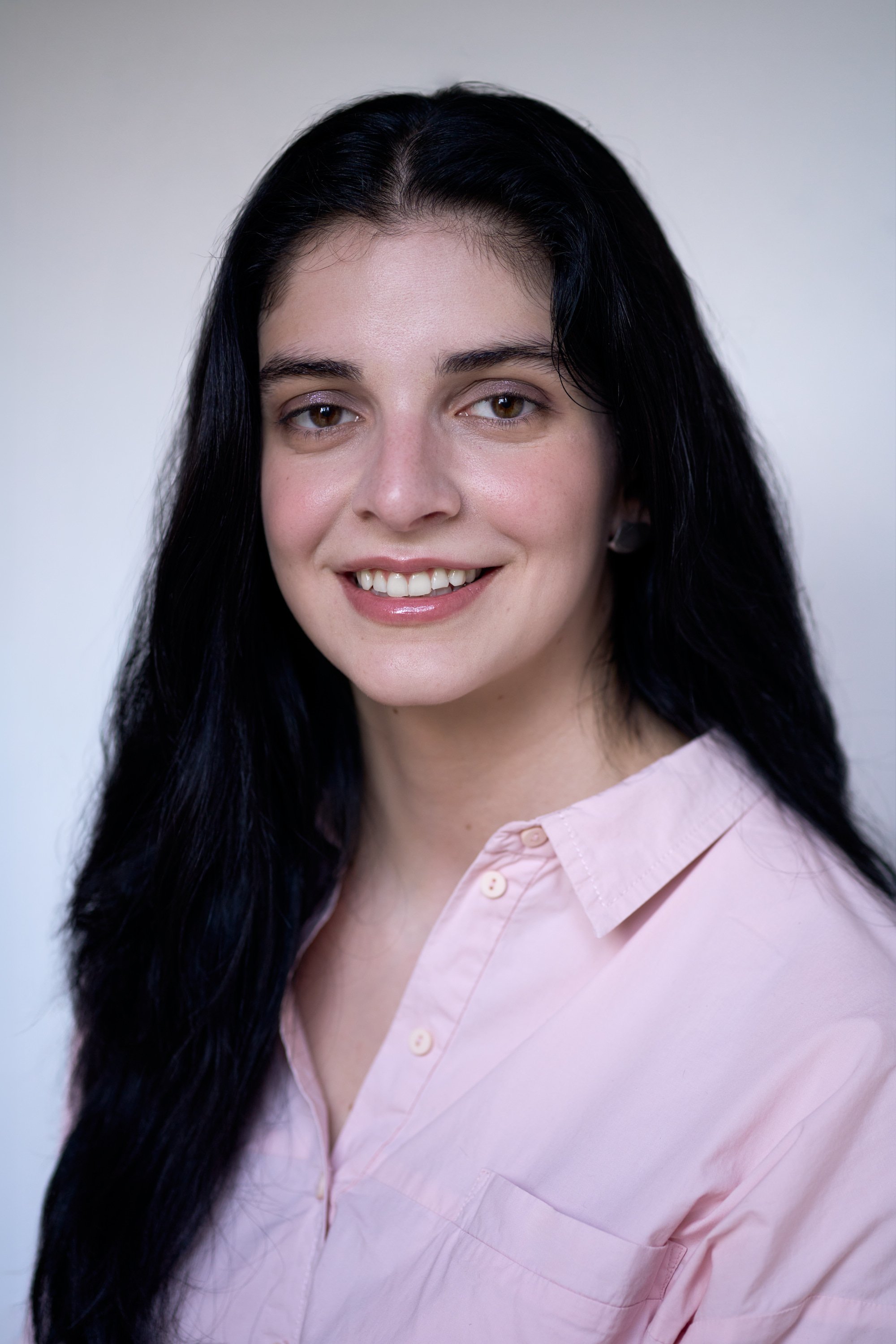 A young woman with long black hair wearing a light pink button-up shirt, smiling against a plain light background.