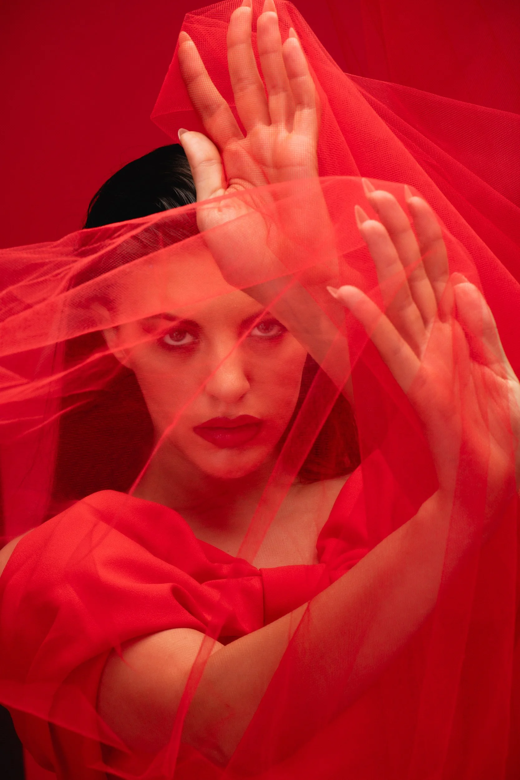 Woman with dark hair and red lipstick behind red sheer fabric, making a dramatic gesture with her hands against a red background.