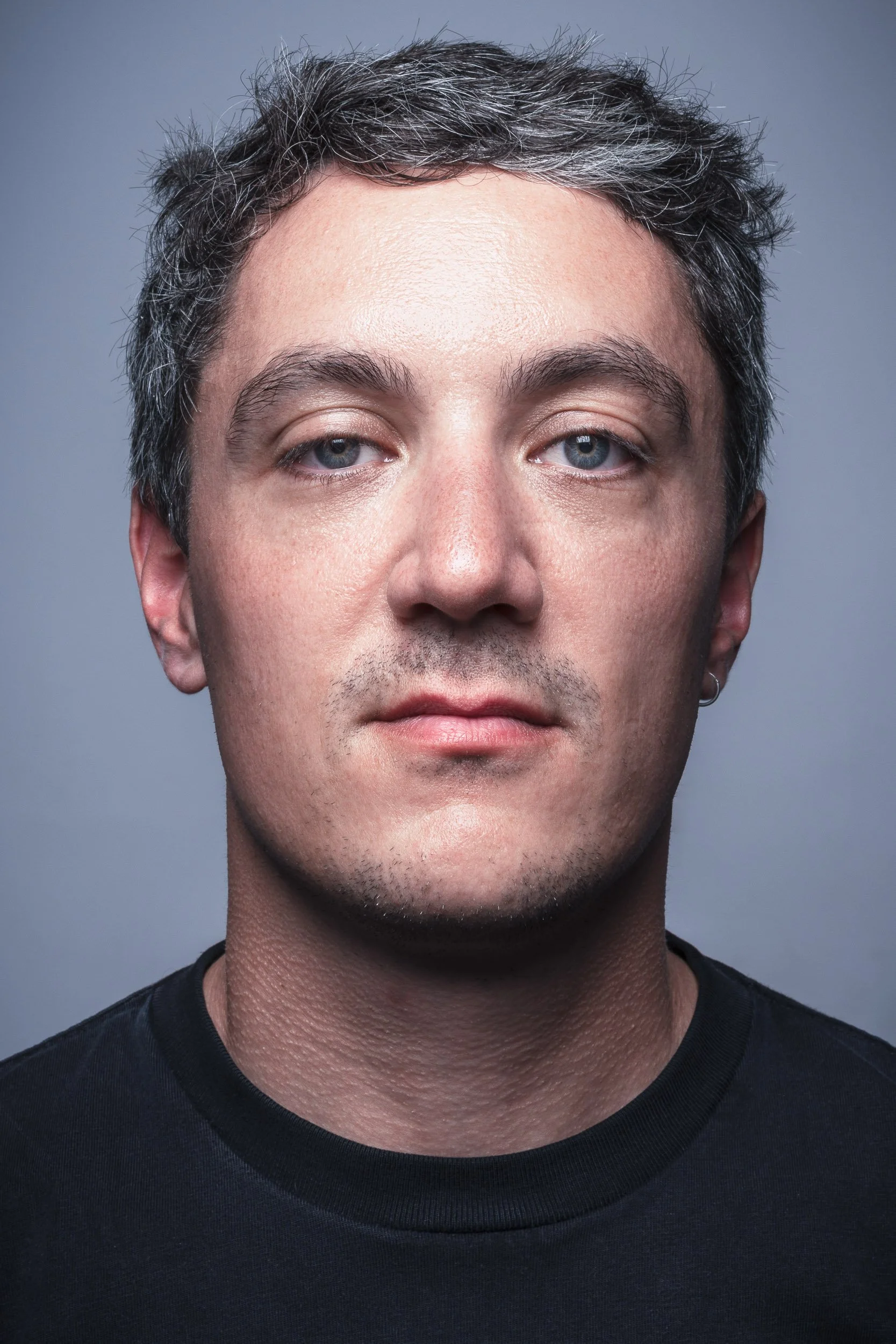 Close-up of a man with short, grayish hair and blue eyes, wearing a black shirt against a gray background.