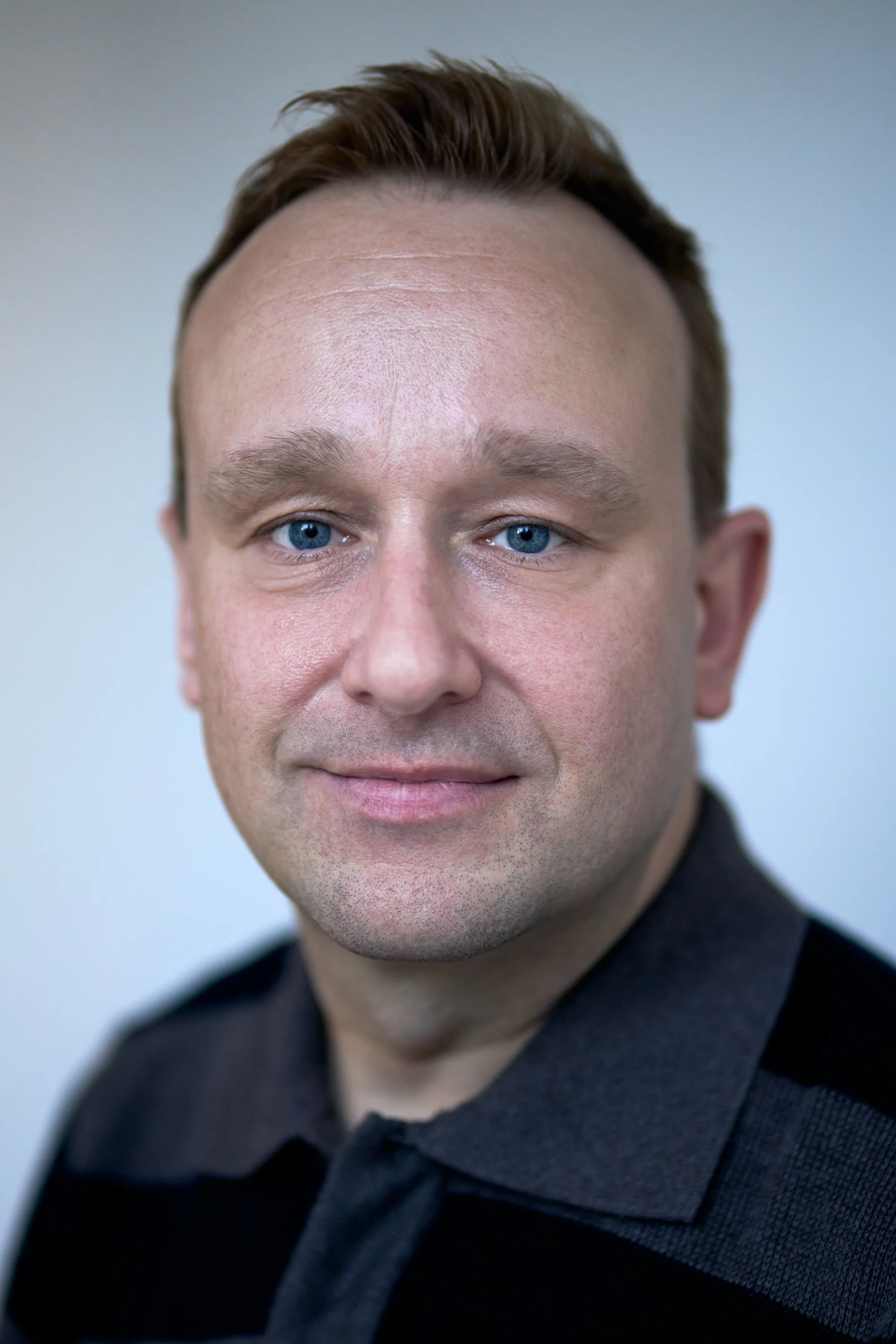Close-up portrait of a man with blue eyes and short brown hair, wearing a dark shirt, against a plain background.