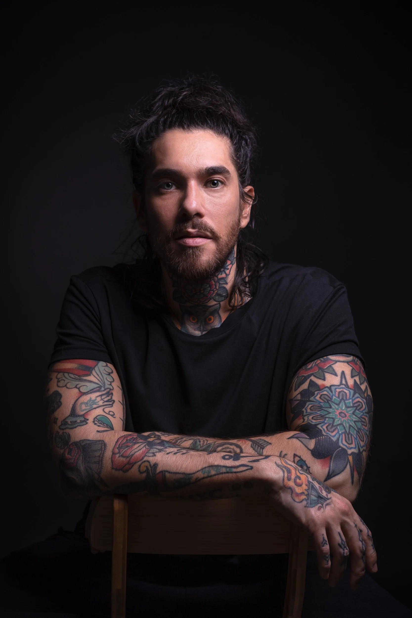 Portrait of a man with dark, curly hair and tattoos on his arms and neck, wearing a black shirt, sitting against a black background.