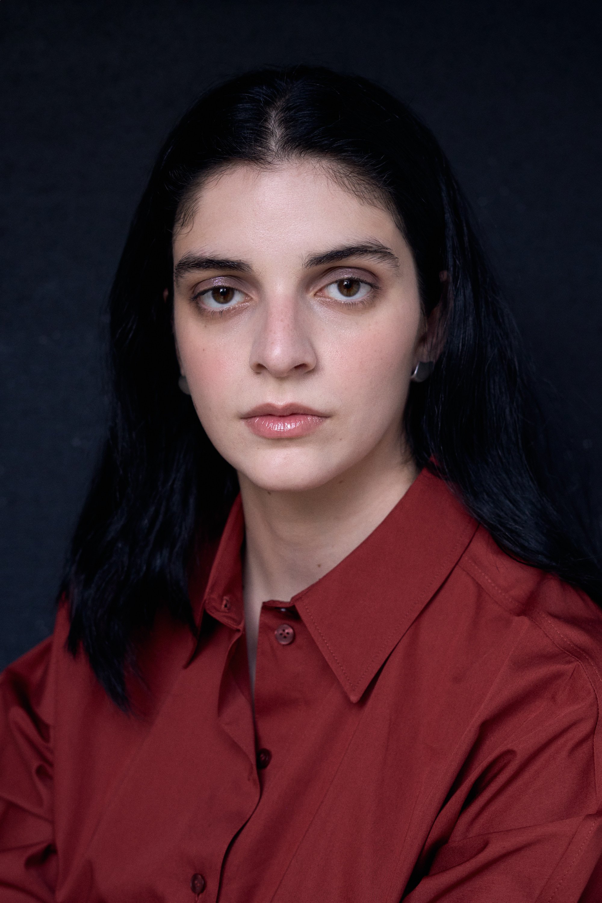 A woman with long dark hair, light skin, and brown eyes, wearing a red button-up shirt, against a dark background.