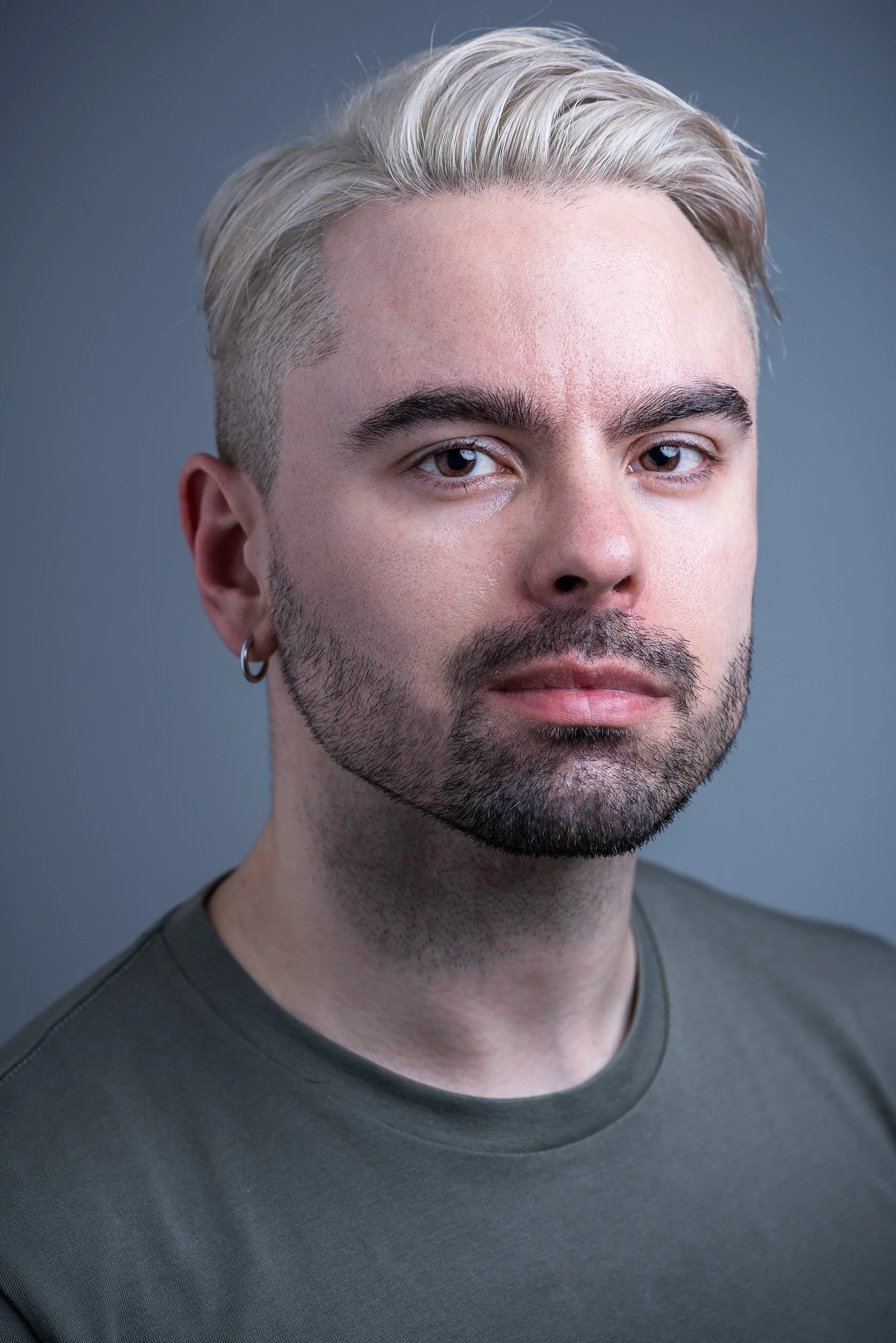 Close-up portrait of a young man with platinum blonde hair, beard, and earrings, wearing a dark green t-shirt, against a plain gray background.