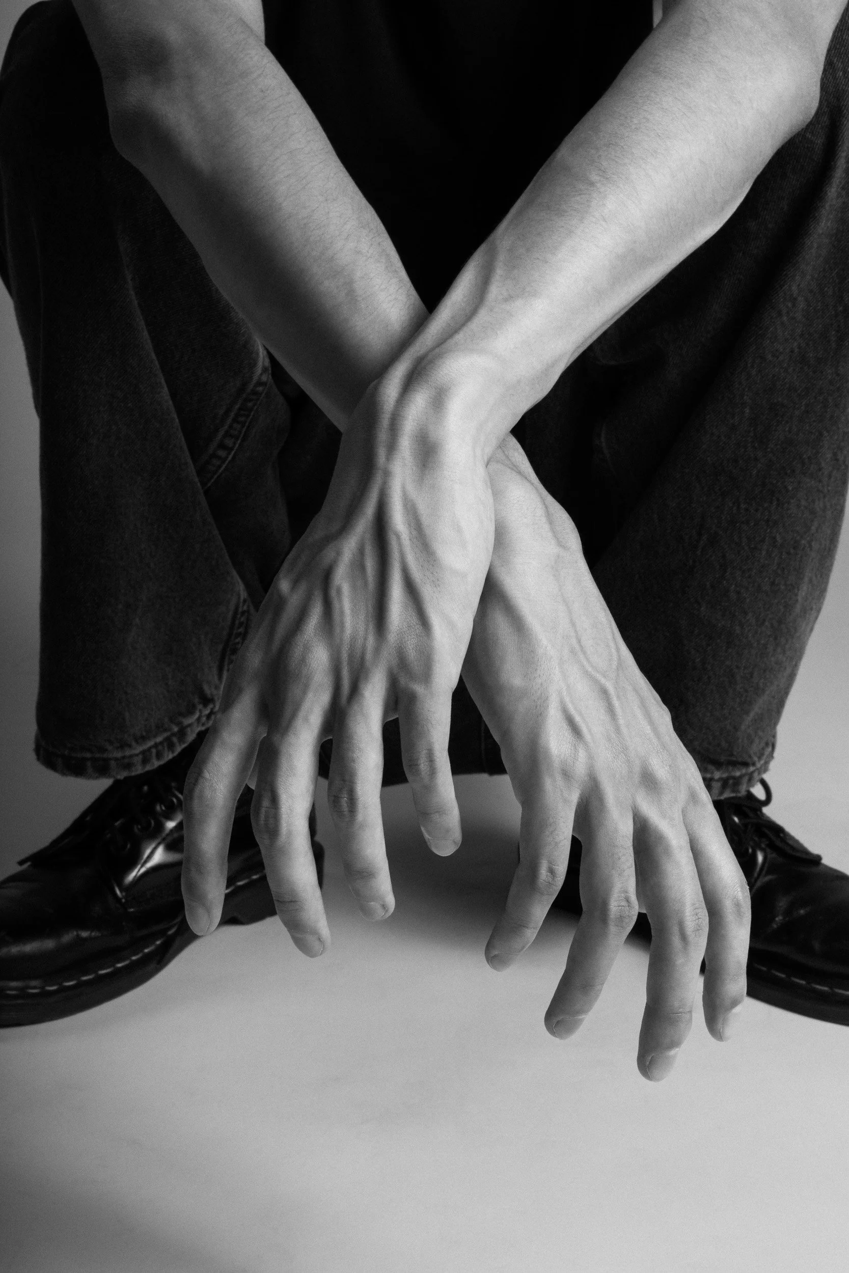 Black and white photo of a person with elongated hands, crouching with hands touching the ground, wearing dark jeans and black shoes.