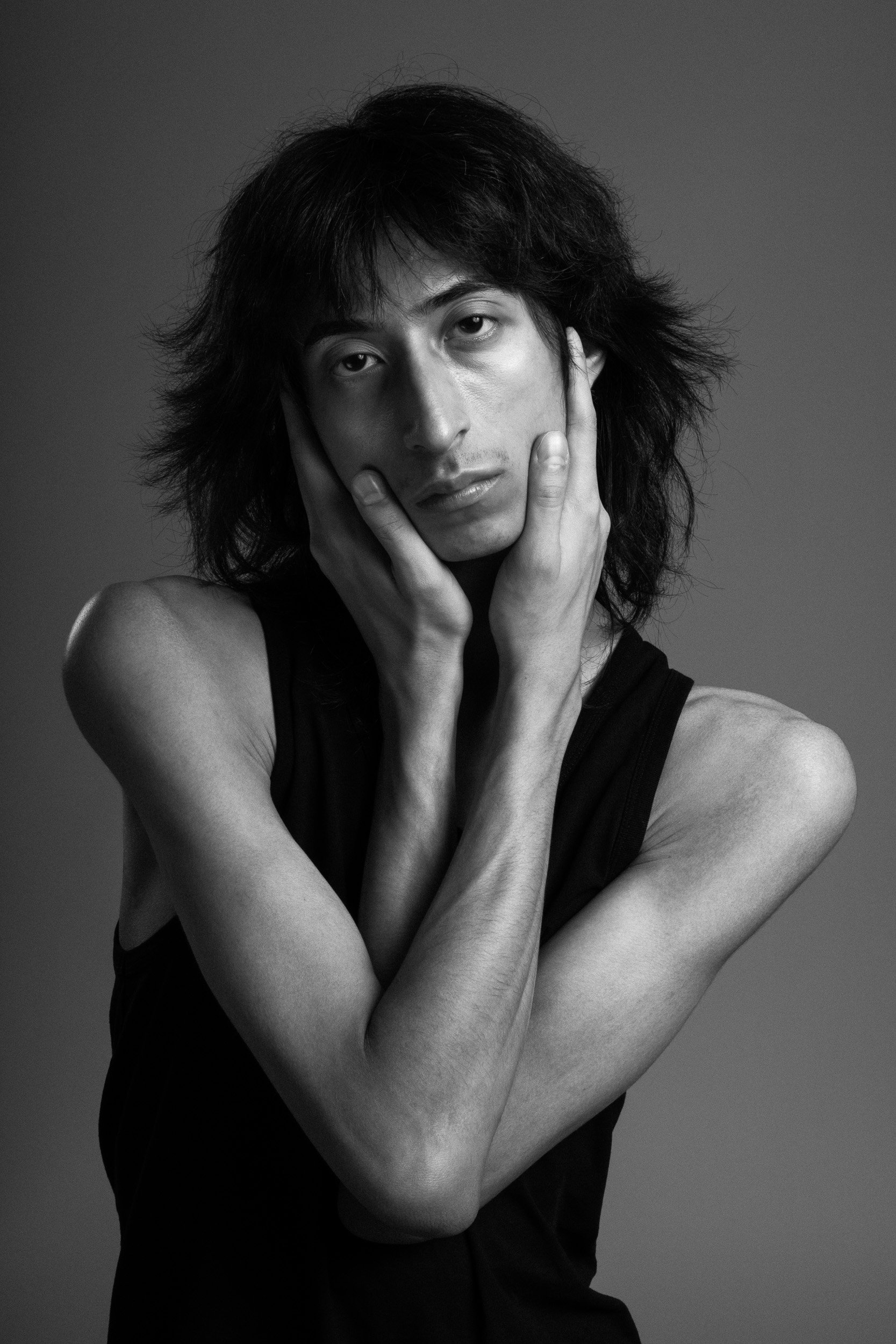 Black and white portrait of a person with shoulder-length hair, touching their face with both hands, wearing a sleeveless top, against a plain background.