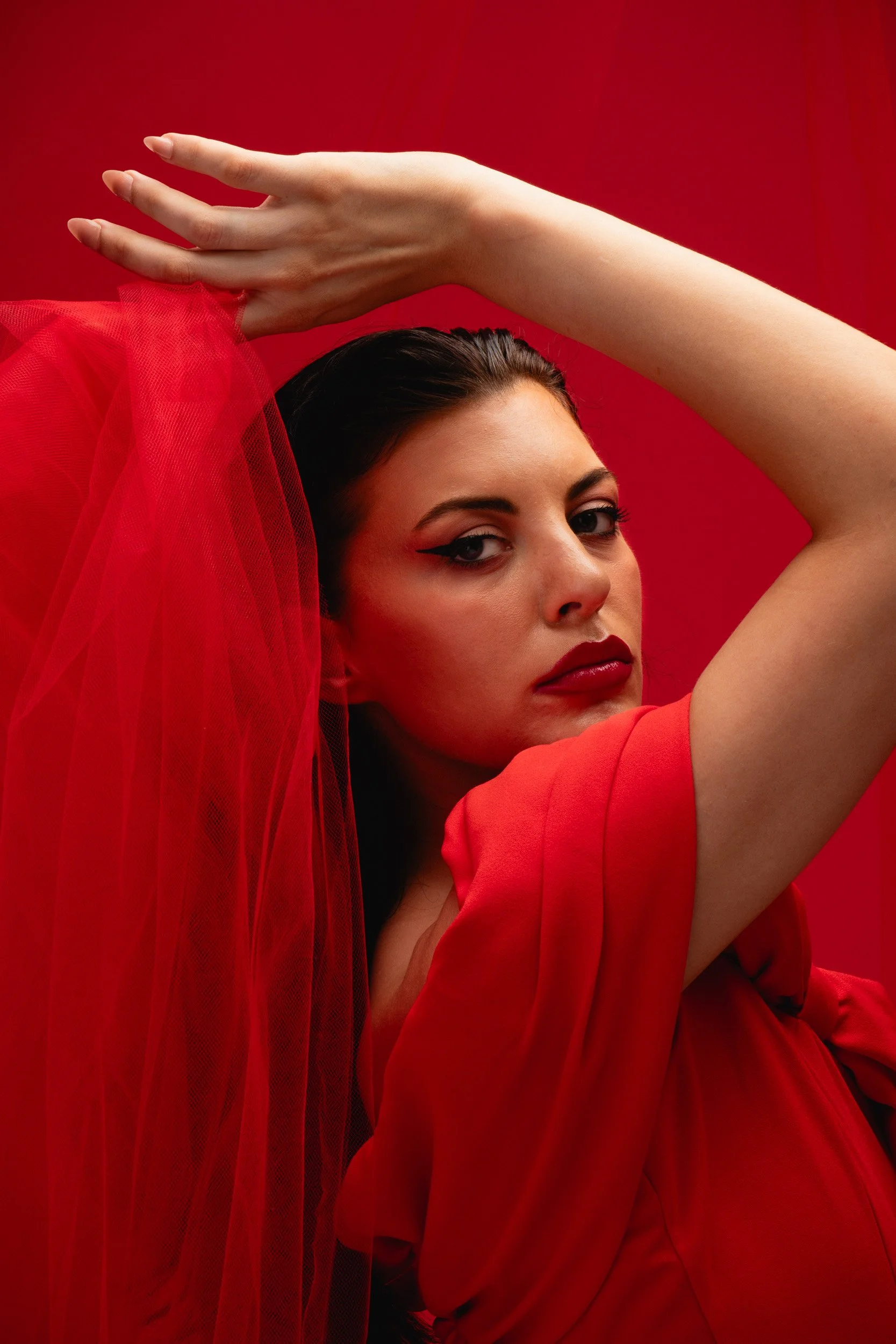 A woman with dark hair and makeup poses against a red background, wearing a red dress and a red veil, with her hand resting above her head.