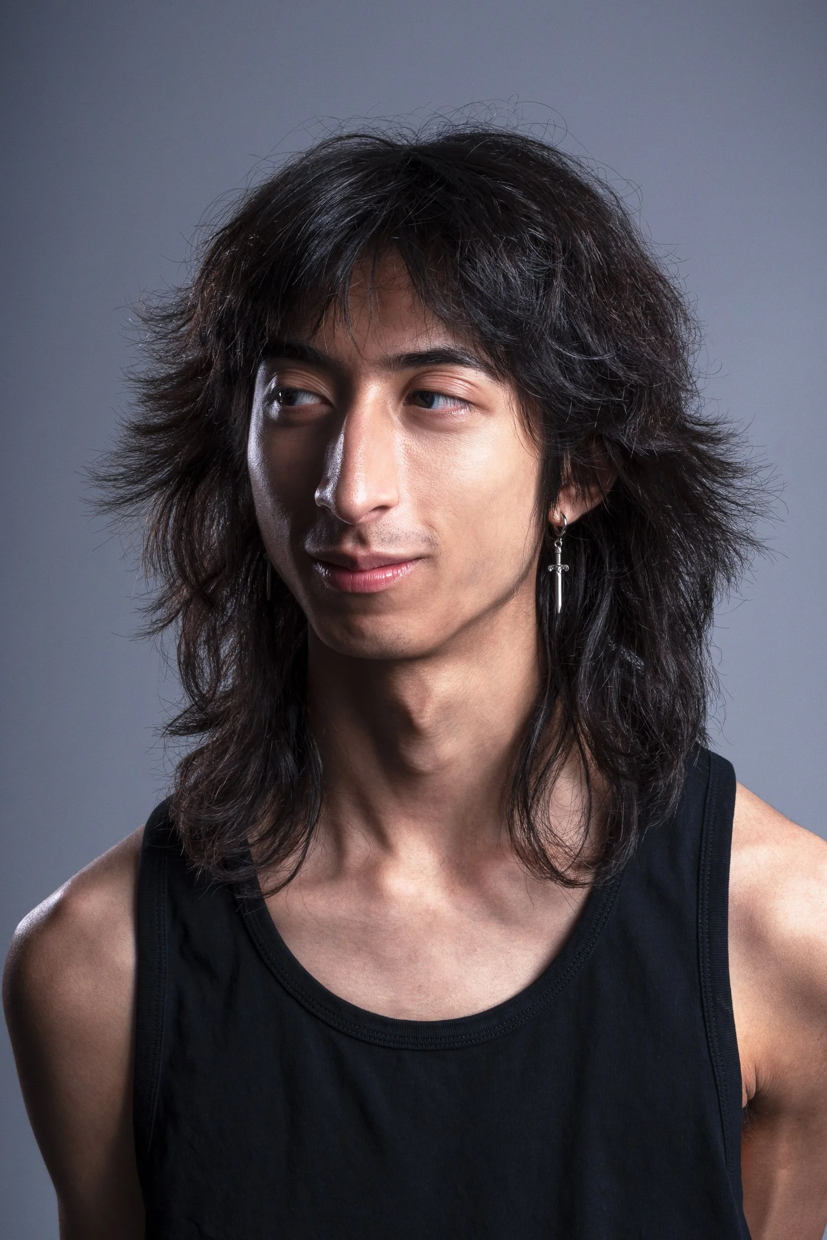 A young man with shoulder-length, wavy dark hair, wearing a black sleeveless top and an earring with a cross. He is looking slightly to his left against a plain gray background.