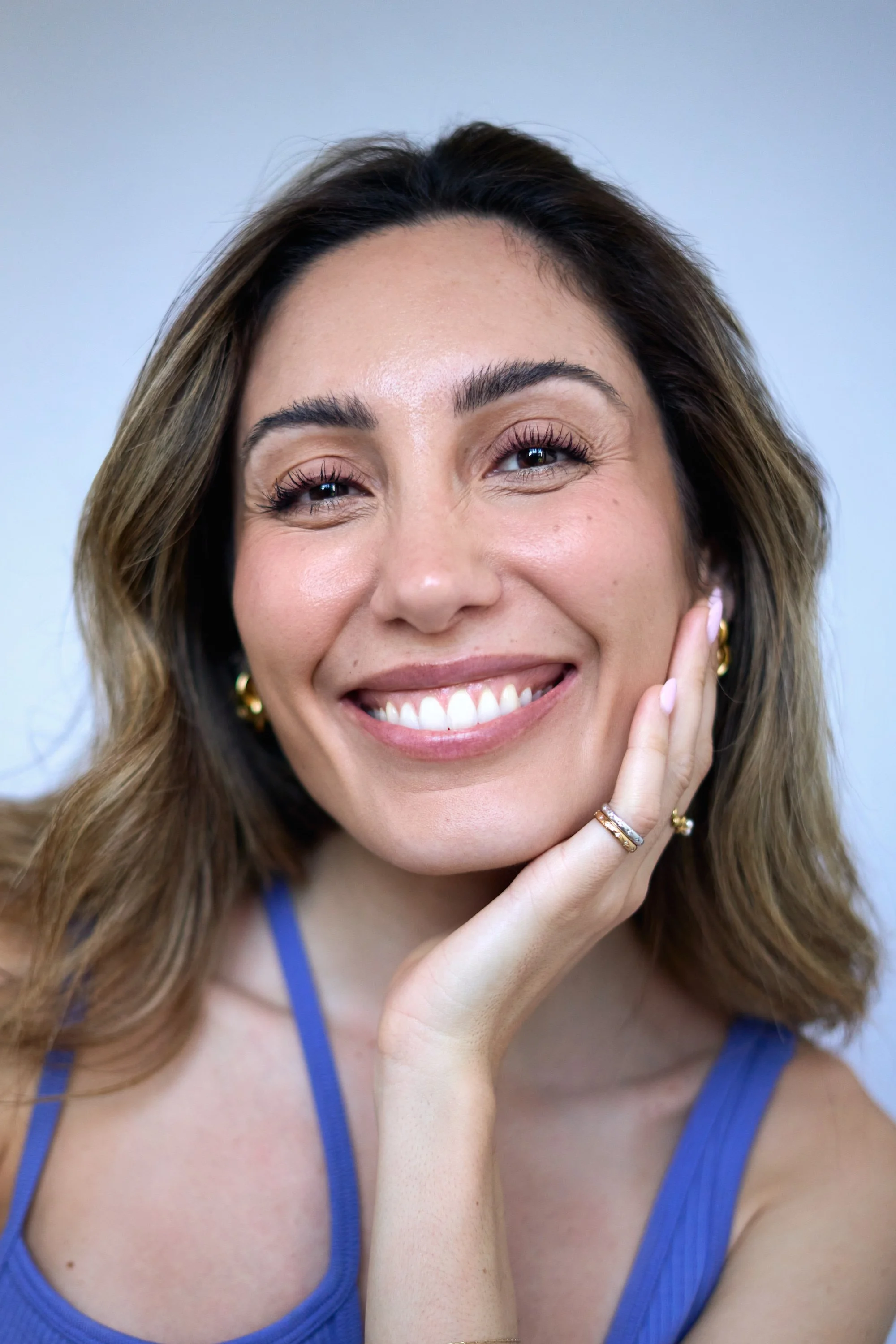 A woman with light brown hair, wearing a blue tank top and gold jewelry, smiling and resting her face on her hand.