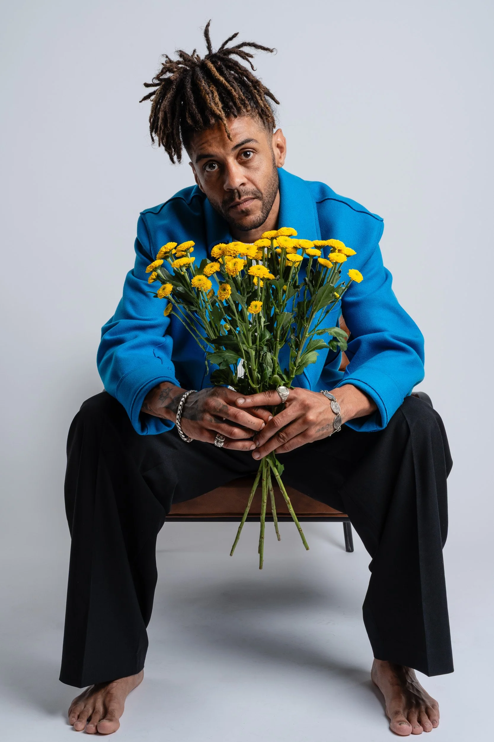 A man with dreadlocks wearing a blue jacket and black pants, sitting barefoot on a chair, holding a large bouquet of yellow flowers, looking directly at the camera with a neutral expression.