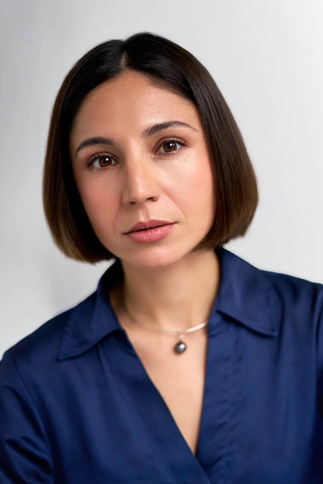 A woman with short brown hair wearing a navy blue blouse and a necklace with a black pendant, looking directly at the camera with a neutral expression.