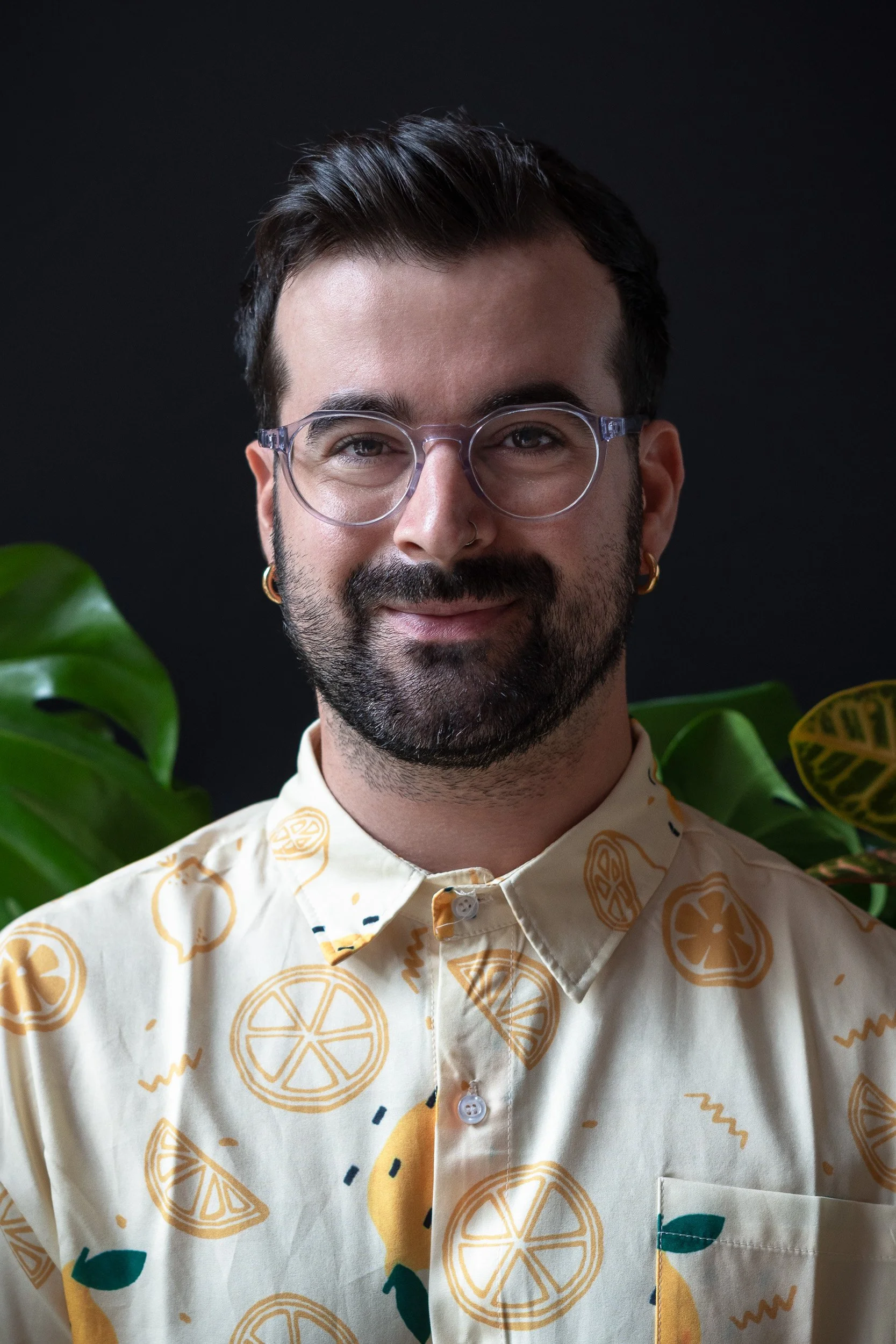 Portrait of a man with glasses, beard, and earrings, wearing a lemon-patterned shirt, standing in front of green plants against a dark background.