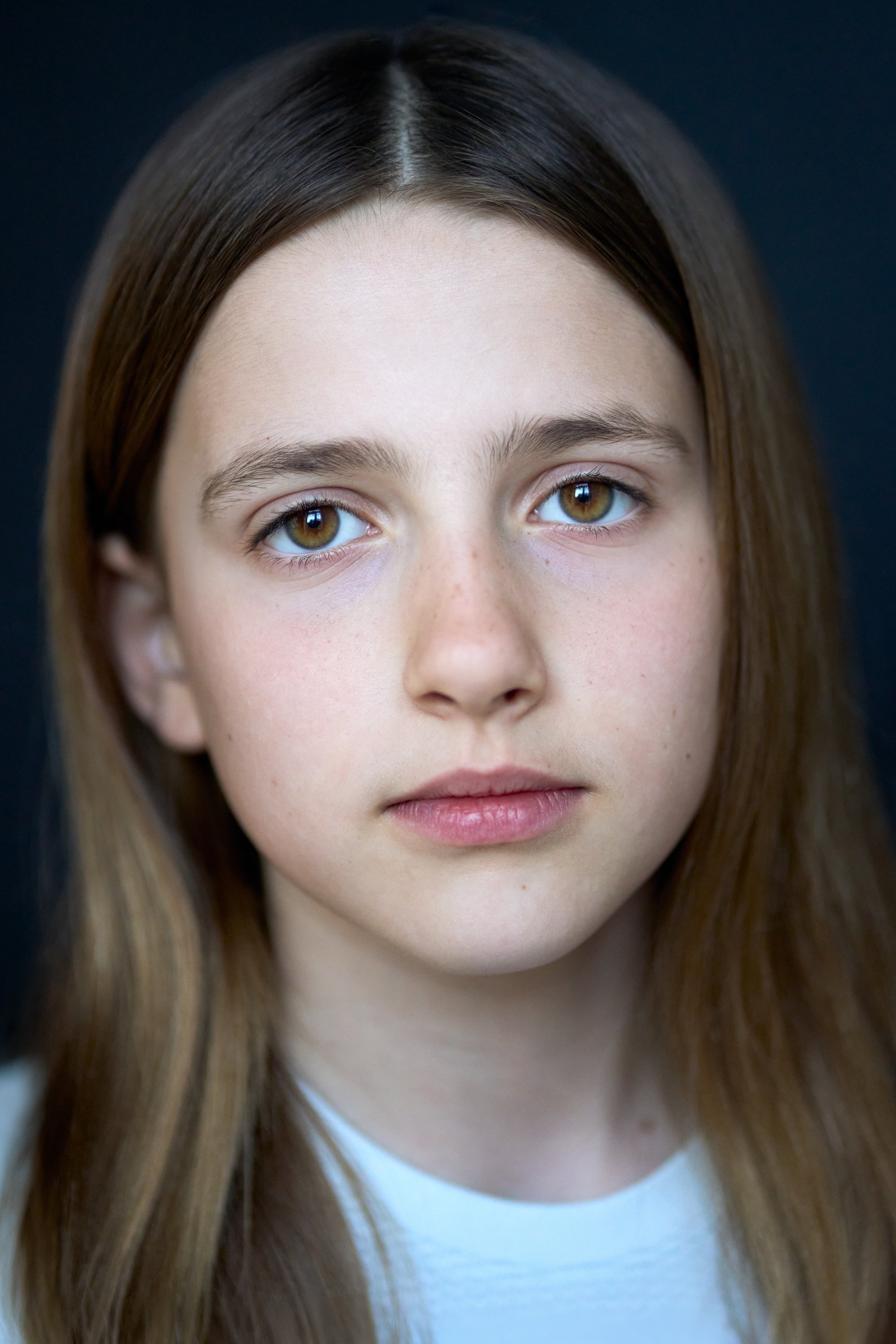 Close-up portrait of a young girl with long brown hair and hazel eyes, looking directly at the camera against a dark background.