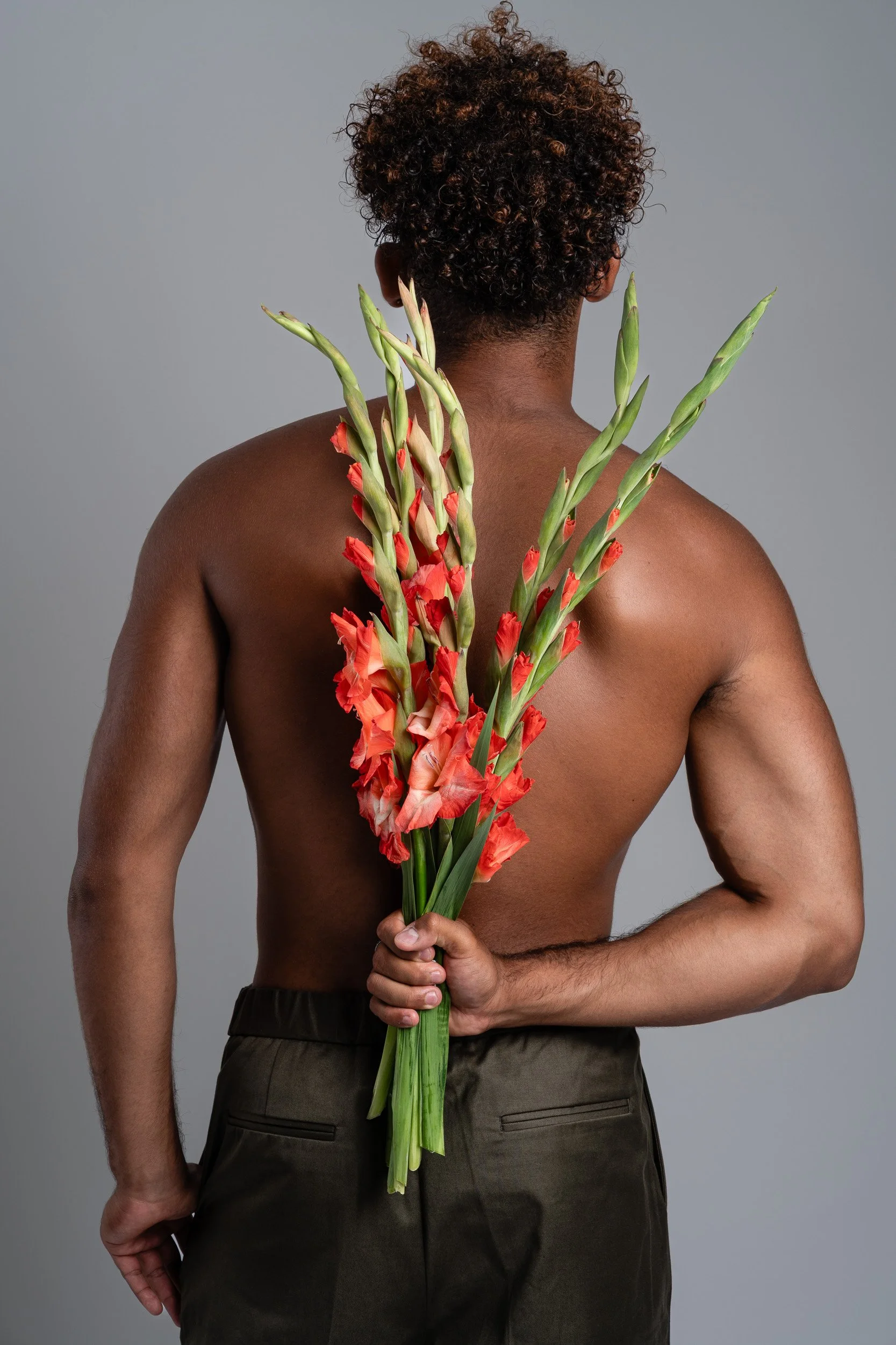 A shirtless person with curly hair holding a bouquet of orange gladiolus flowers behind their back, against a plain gray background.