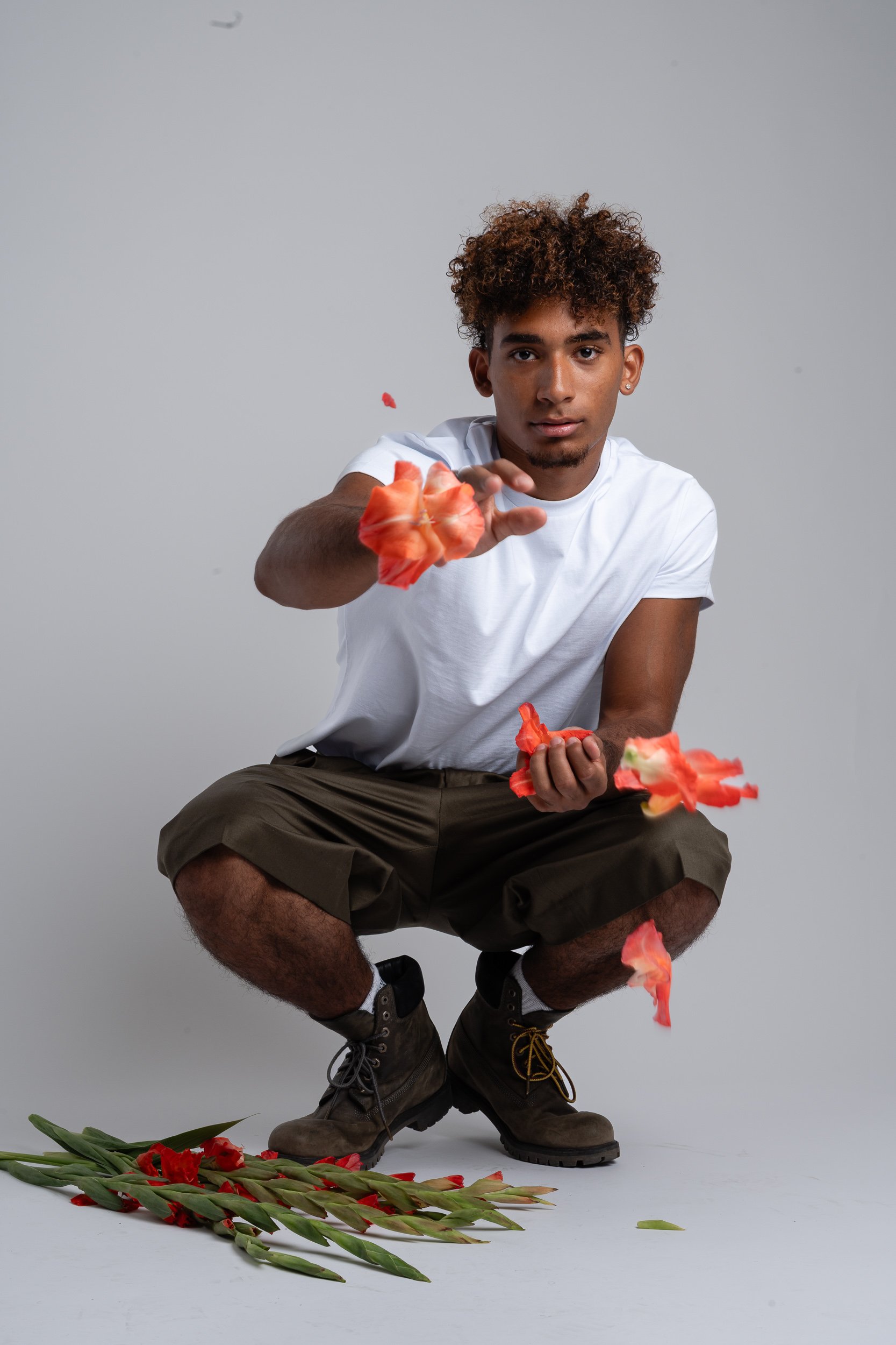 Young man crouching, throwing flower petals, with more petals and lilies on the floor, against a plain gray background.