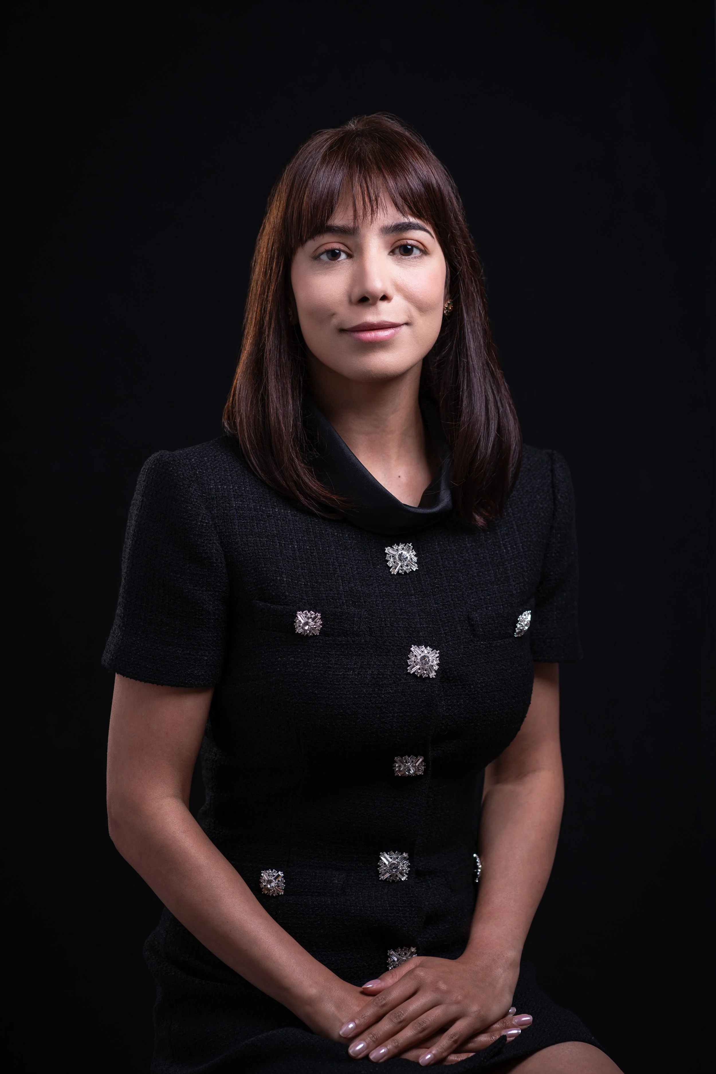 A woman with shoulder-length brown hair, wearing a black dress with decorative silver buttons, sitting against a black background.