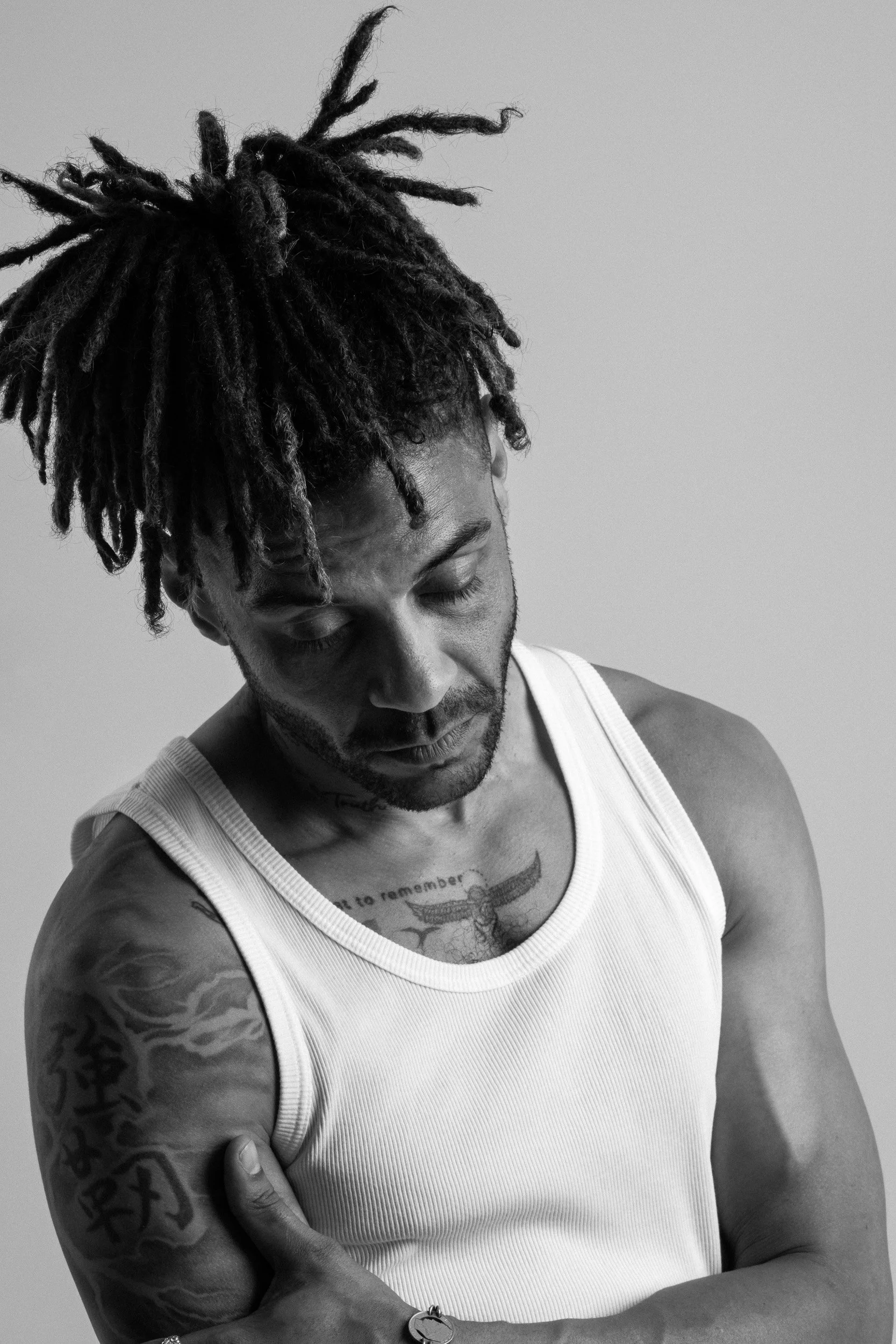 A man with dreadlocks, wearing a white tank top, looking down with eyes closed, standing against a plain background.