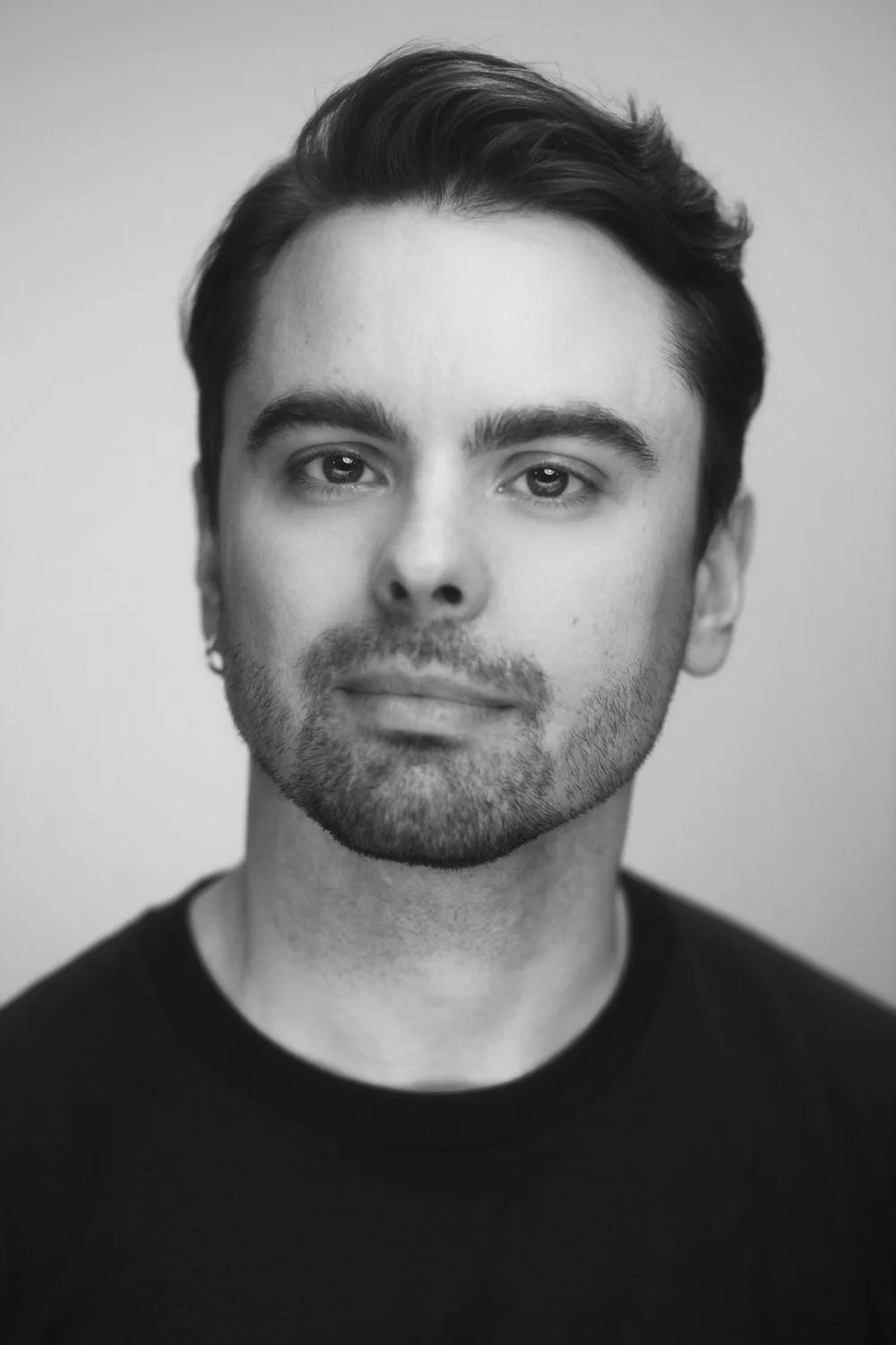Black and white portrait of a young man with dark hair, slight beard, and mustache, wearing a black shirt, looking directly at the camera.
