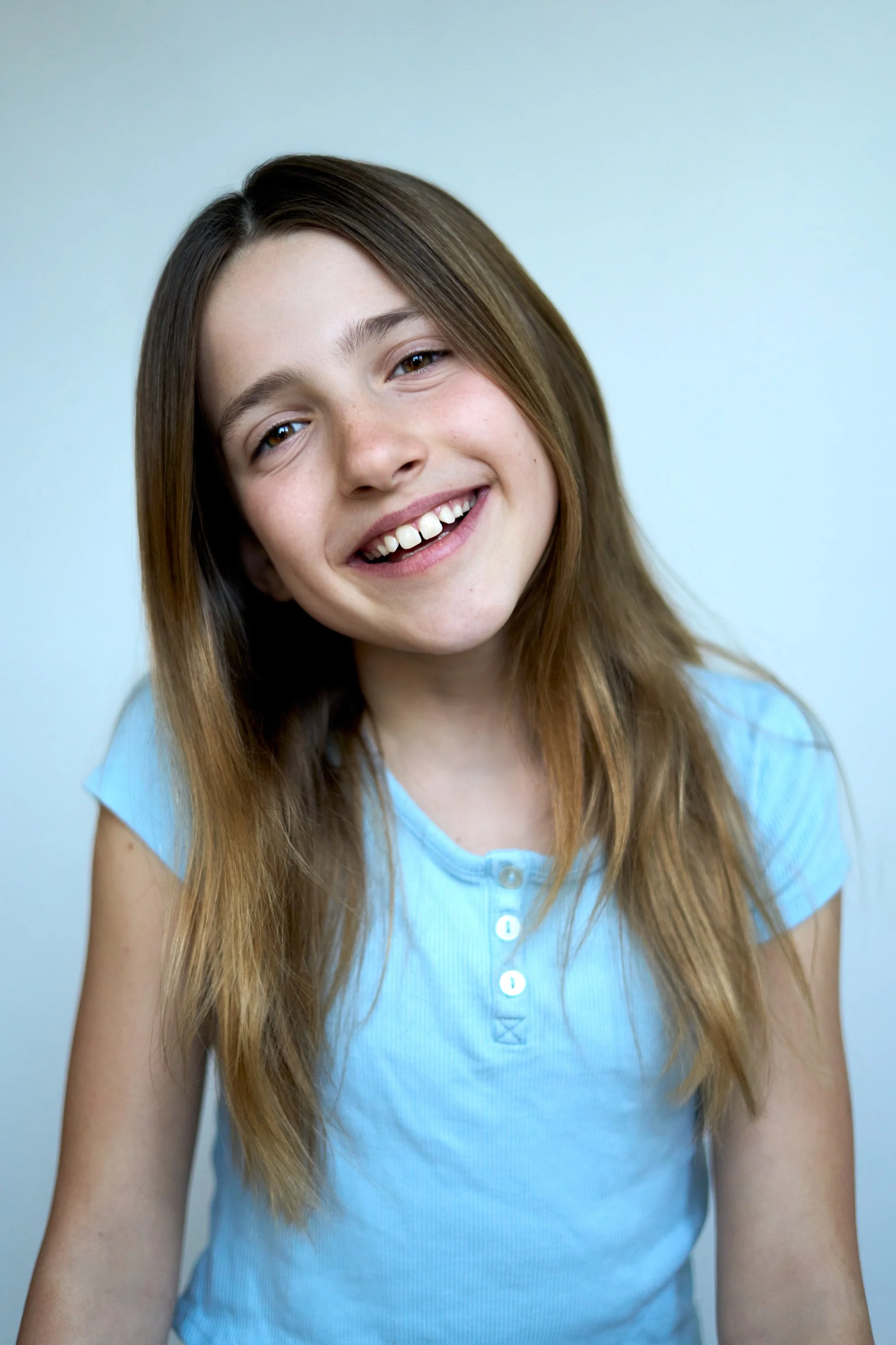 A young girl with long brown hair, smiling, wearing a light blue button-up shirt, against a plain light background.