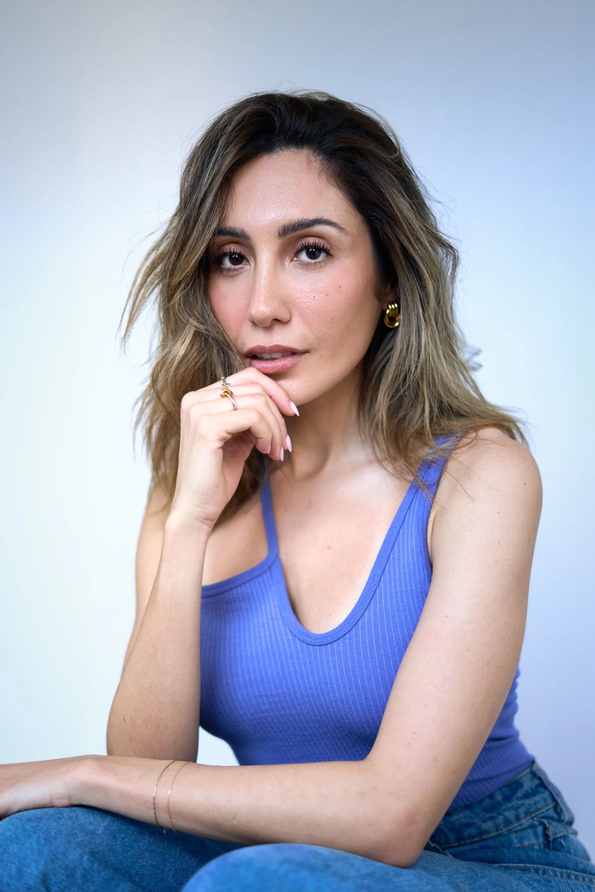 A woman with shoulder-length wavy brown hair, wearing a sleeveless blue top, sitting with her hand near her face, and looking directly at the camera against a plain light background.