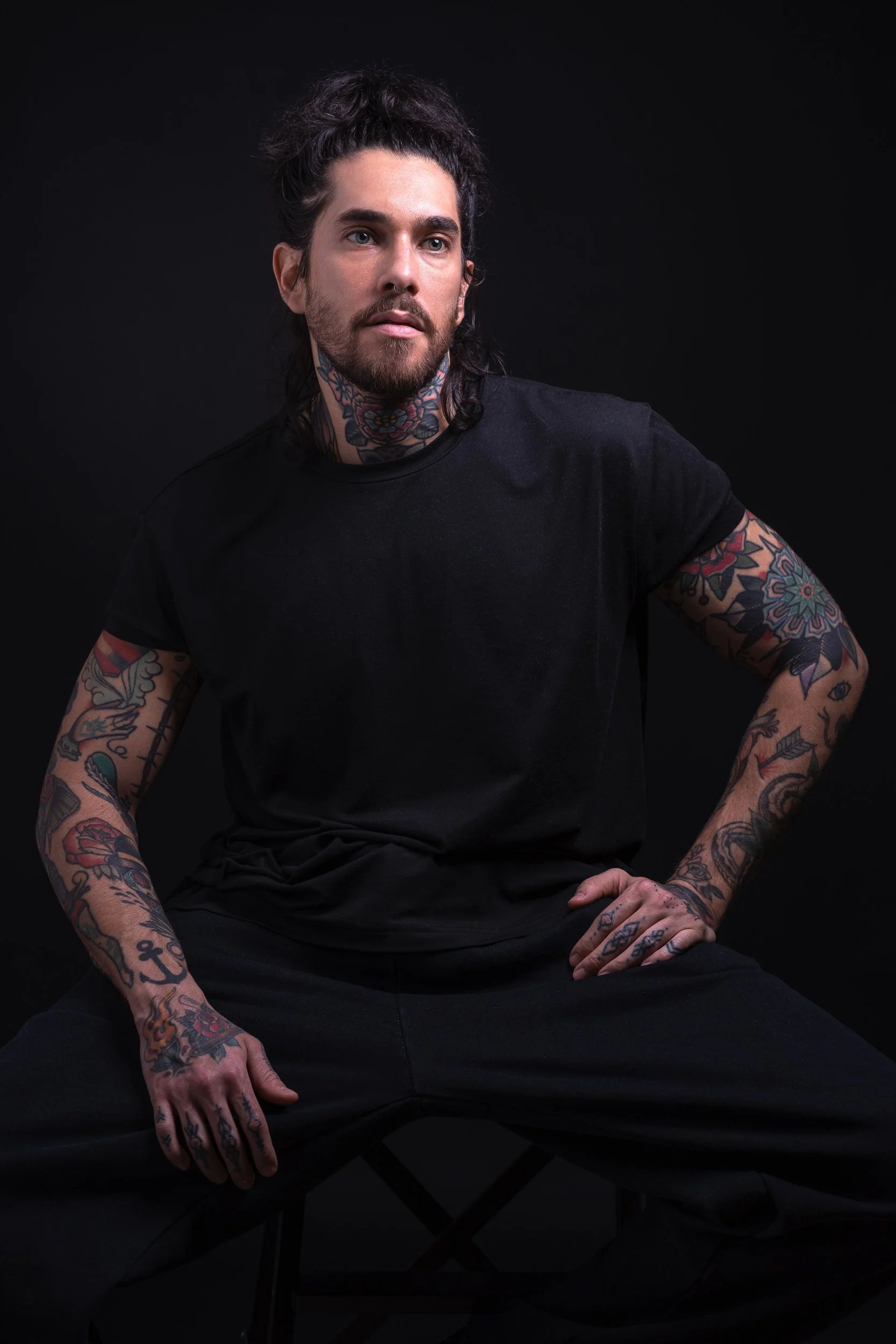 A man with dark hair, beard, and tattoos wearing a black t-shirt and black pants, sitting against a dark background.