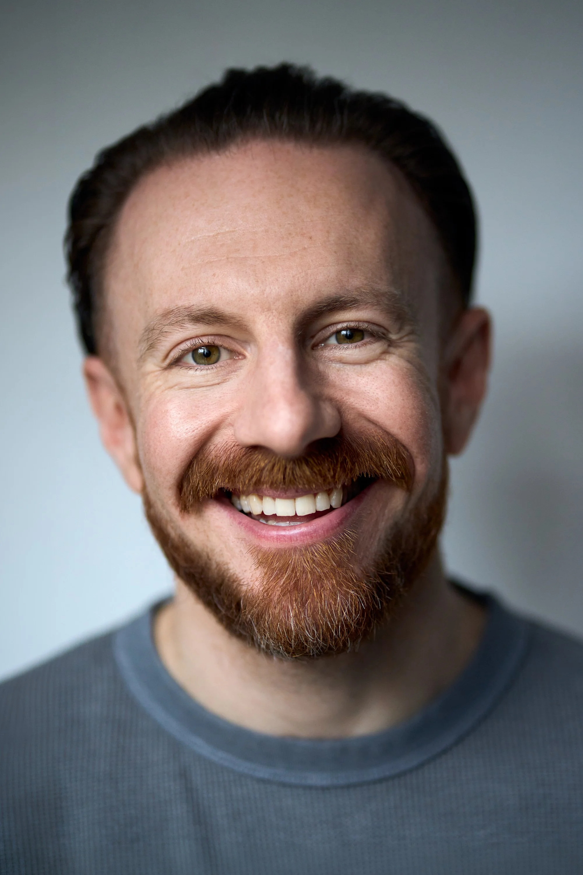 Close-up of smiling man with short dark hair, beard, and green eyes, wearing a gray shirt.