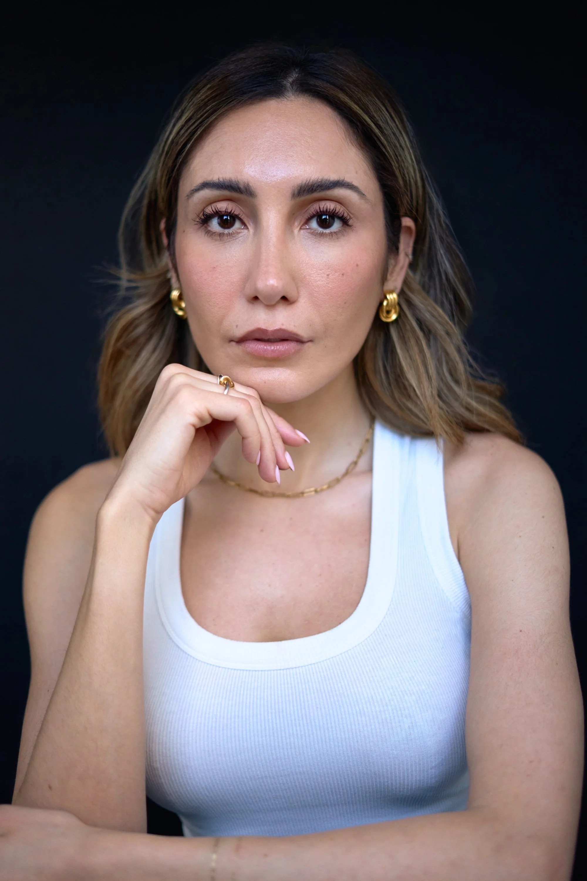 A woman with shoulder-length brown hair, wearing gold jewelry, a white tank top, and a neutral expression, sitting against a dark background.