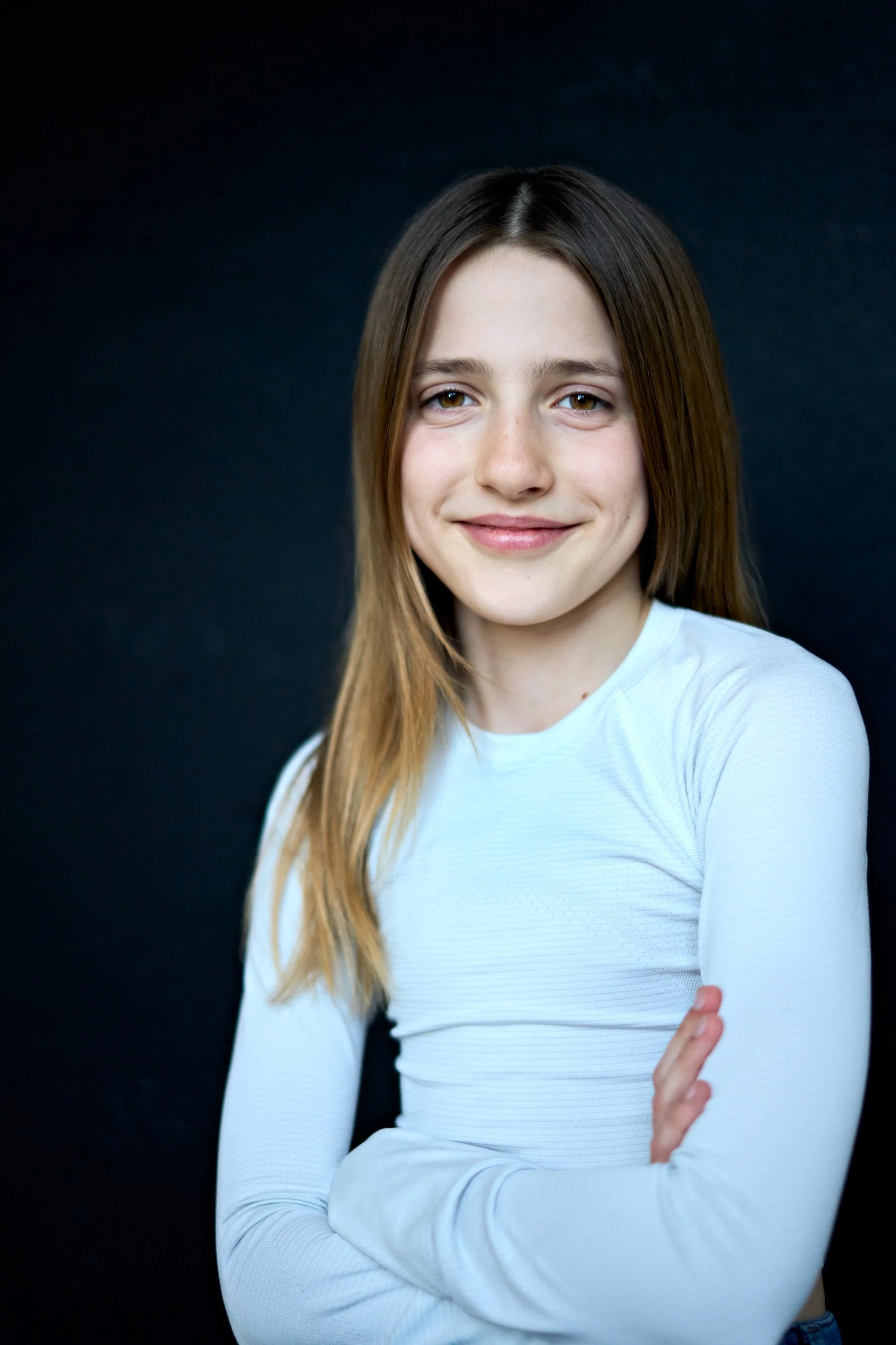 Portrait of a young woman with long brown hair, smiling, wearing a white long-sleeve top, against a dark background.