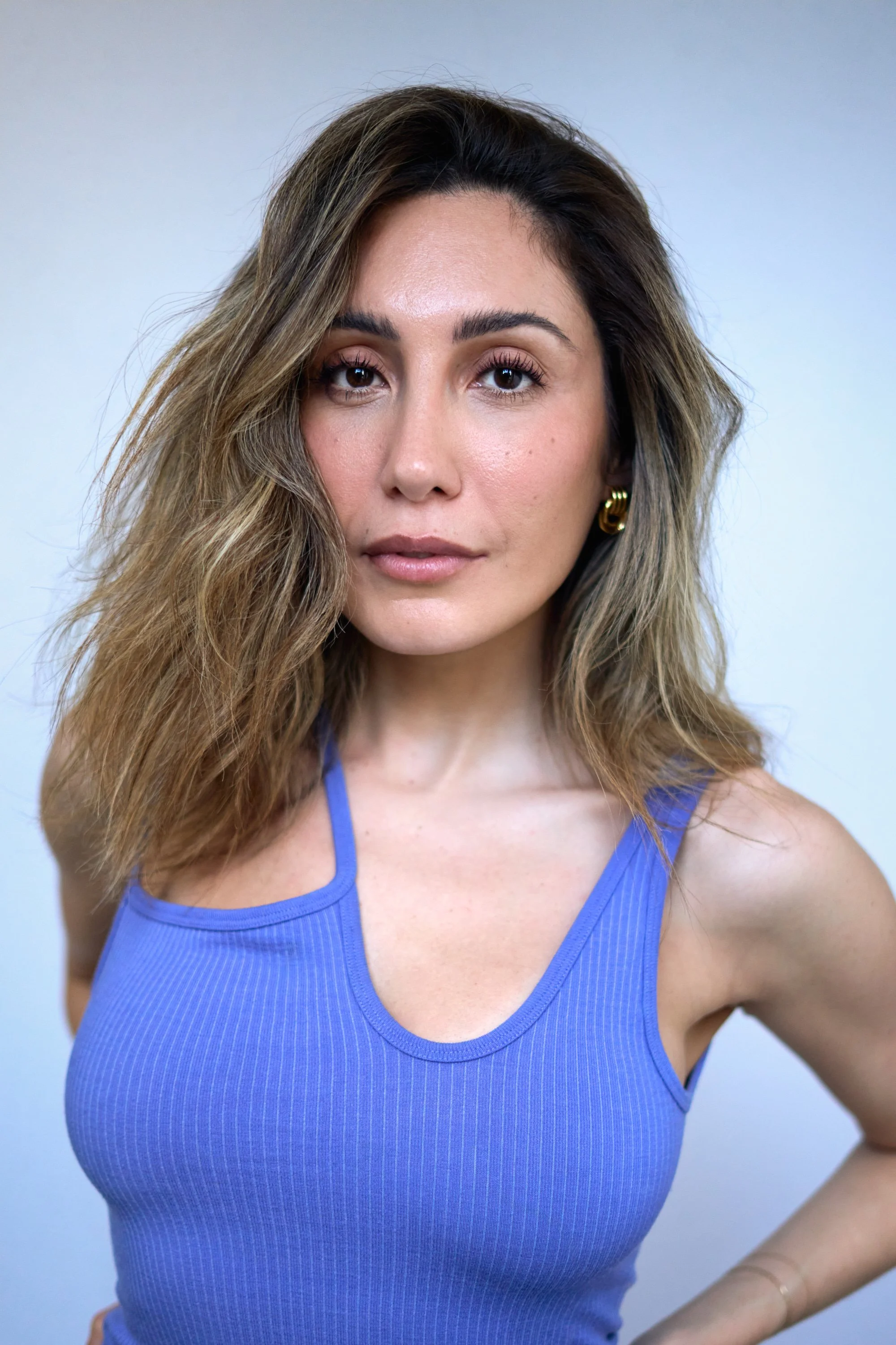 Close-up portrait of a woman with shoulder-length wavy hair wearing a blue sleeveless top with a scoop neckline, standing against a plain light background.