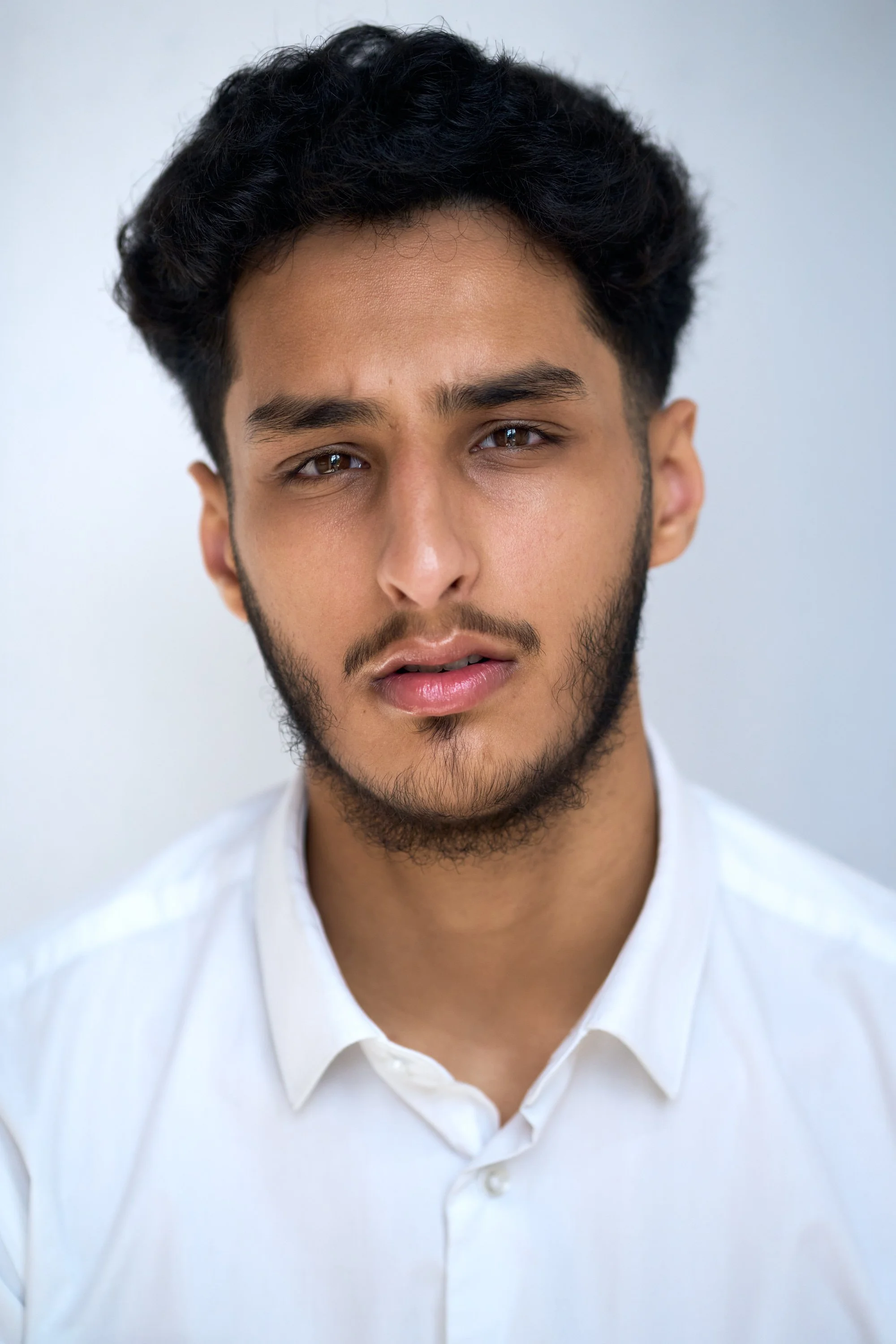 Close-up of a young man with dark, curly hair and light brown skin wearing a white collared shirt, facing the camera with a neutral expression.