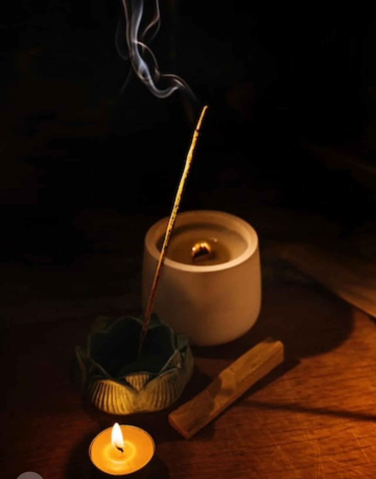 A lit candle, an incense stick burning with smoke, a small wooden object, and a ceramic holder on a wooden surface in darkness.