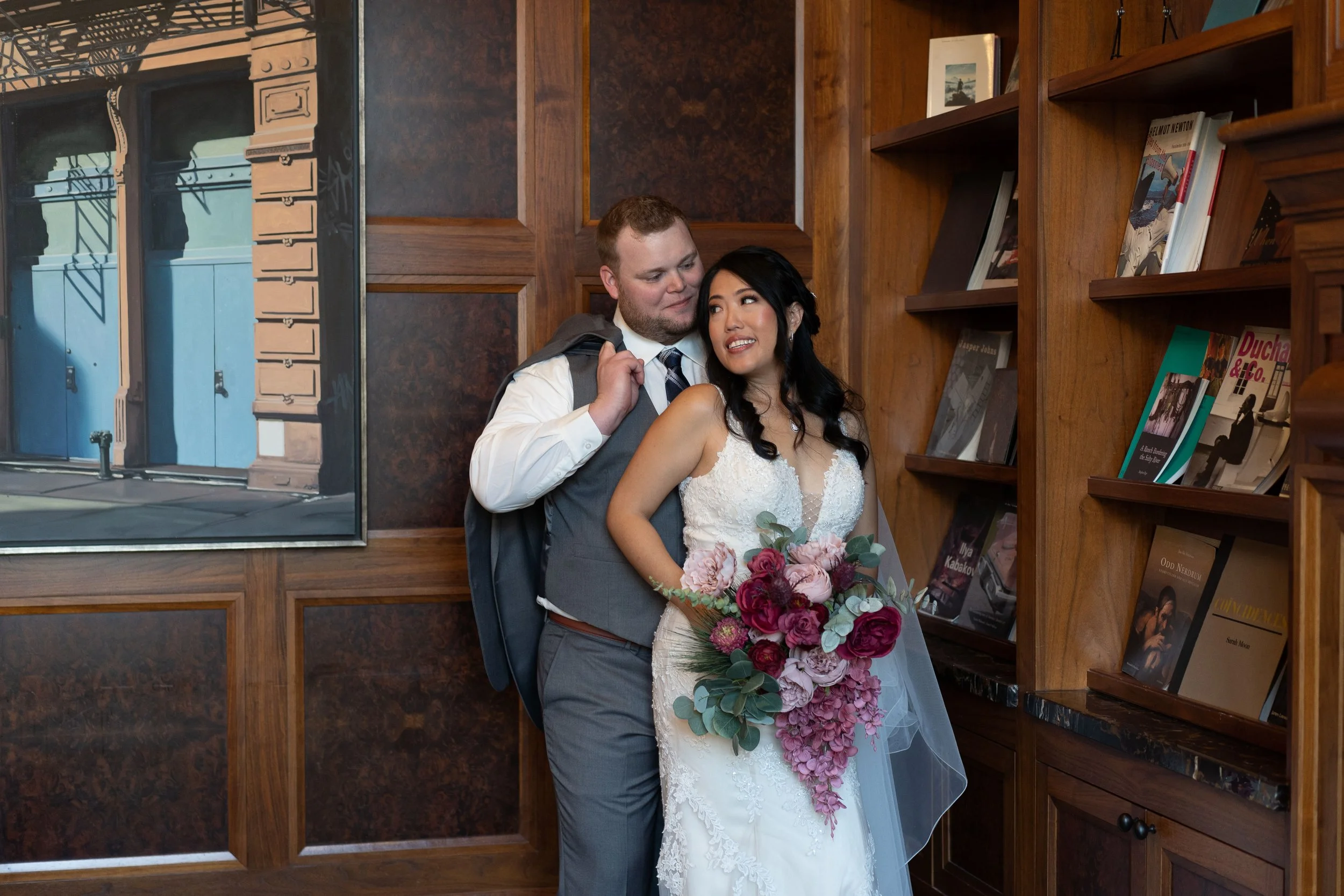 A newlywed couple stands indoors; the groom, with short hair and a beard, in a gray suit and white shirt, carries a backpack over his shoulder, while the bride, with long dark hair, wears a white lace wedding dress and holds a large bouquet of pink a