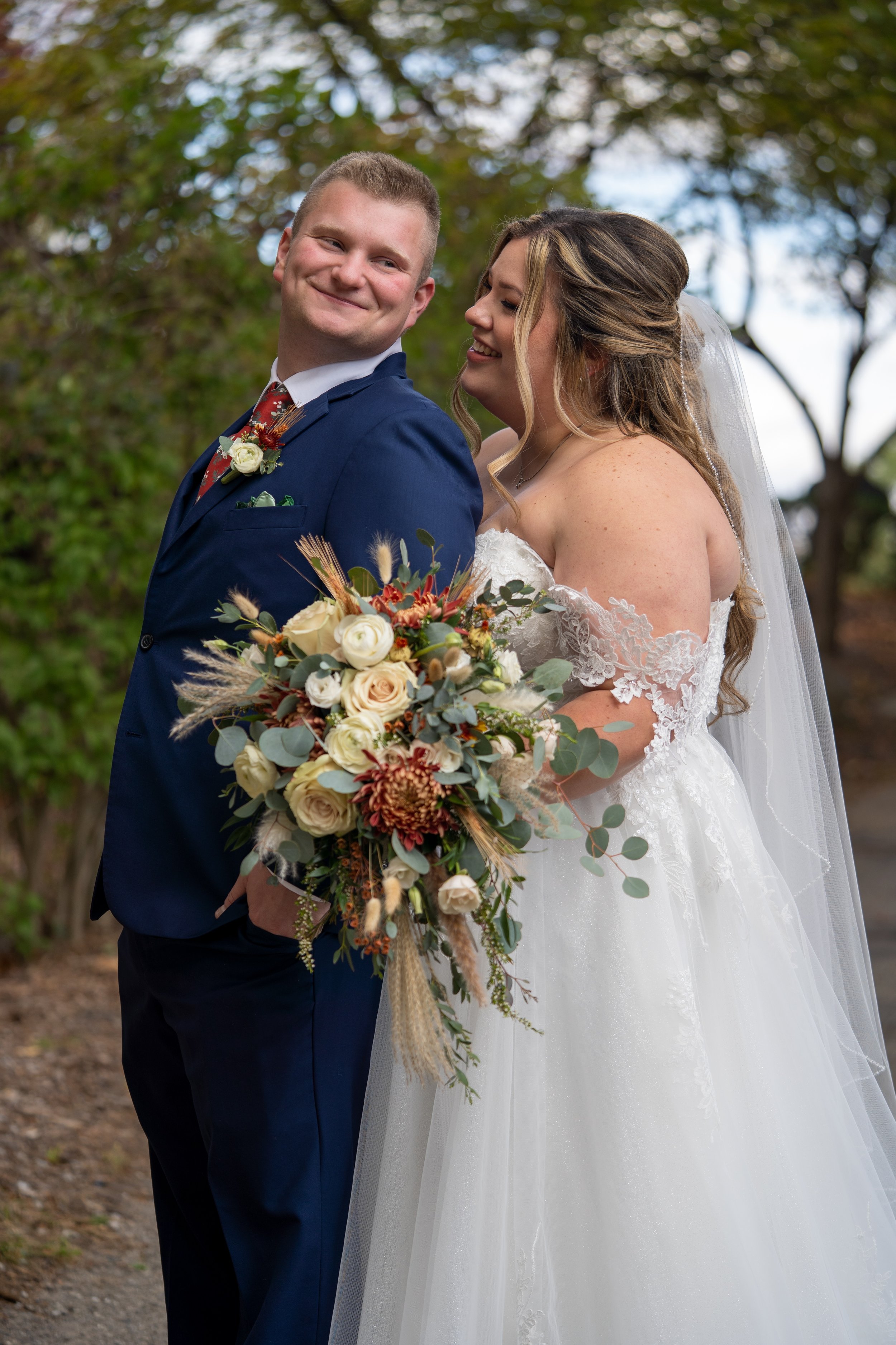 A bride and groom pose outdoors on their wedding day, the bride holding a bouquet of flowers, both smiling and dressed in wedding attire with trees in the background.