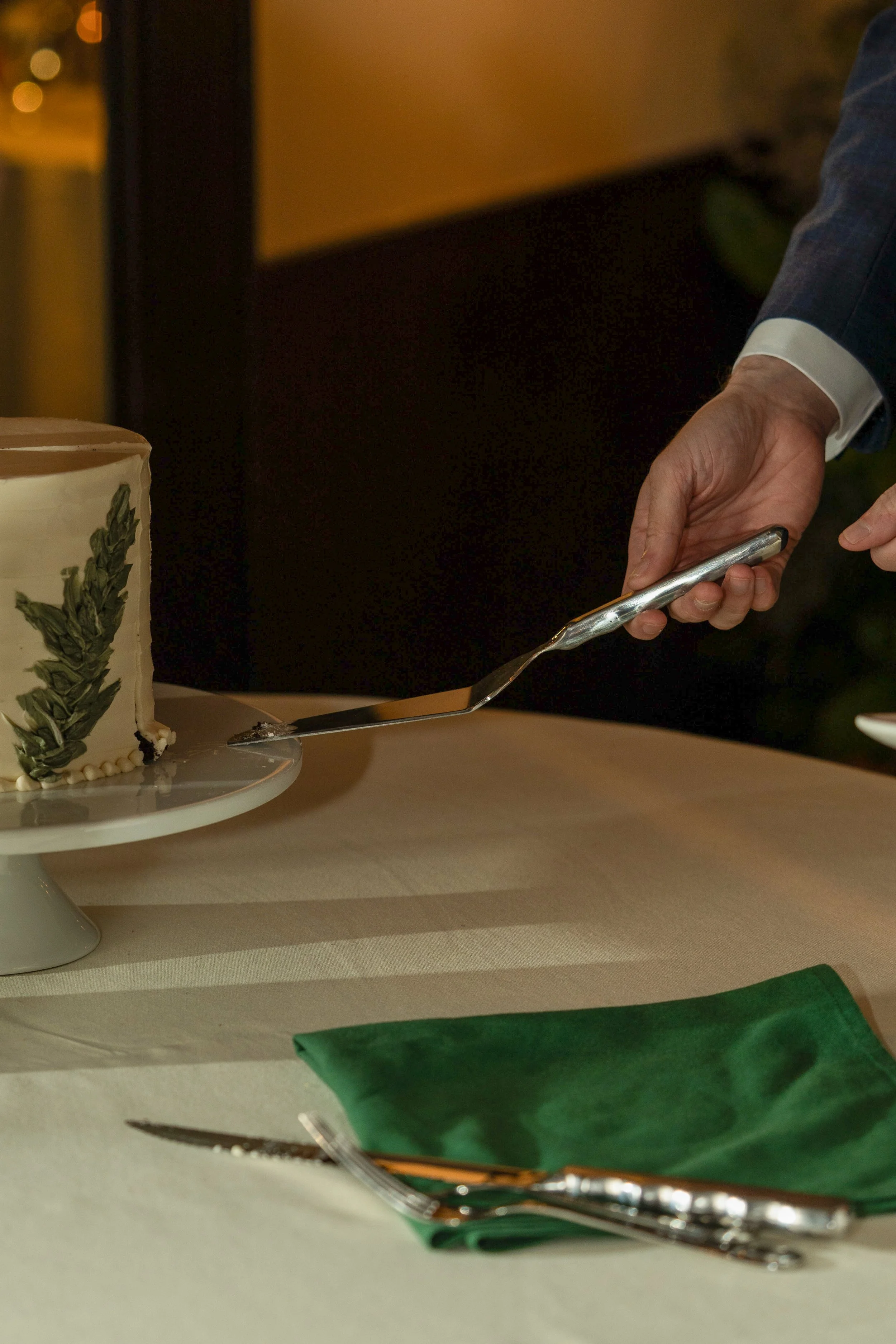 Person in suit cutting a decorated cake with a knife, on a table with a green napkin, a cake server, and a small plate.