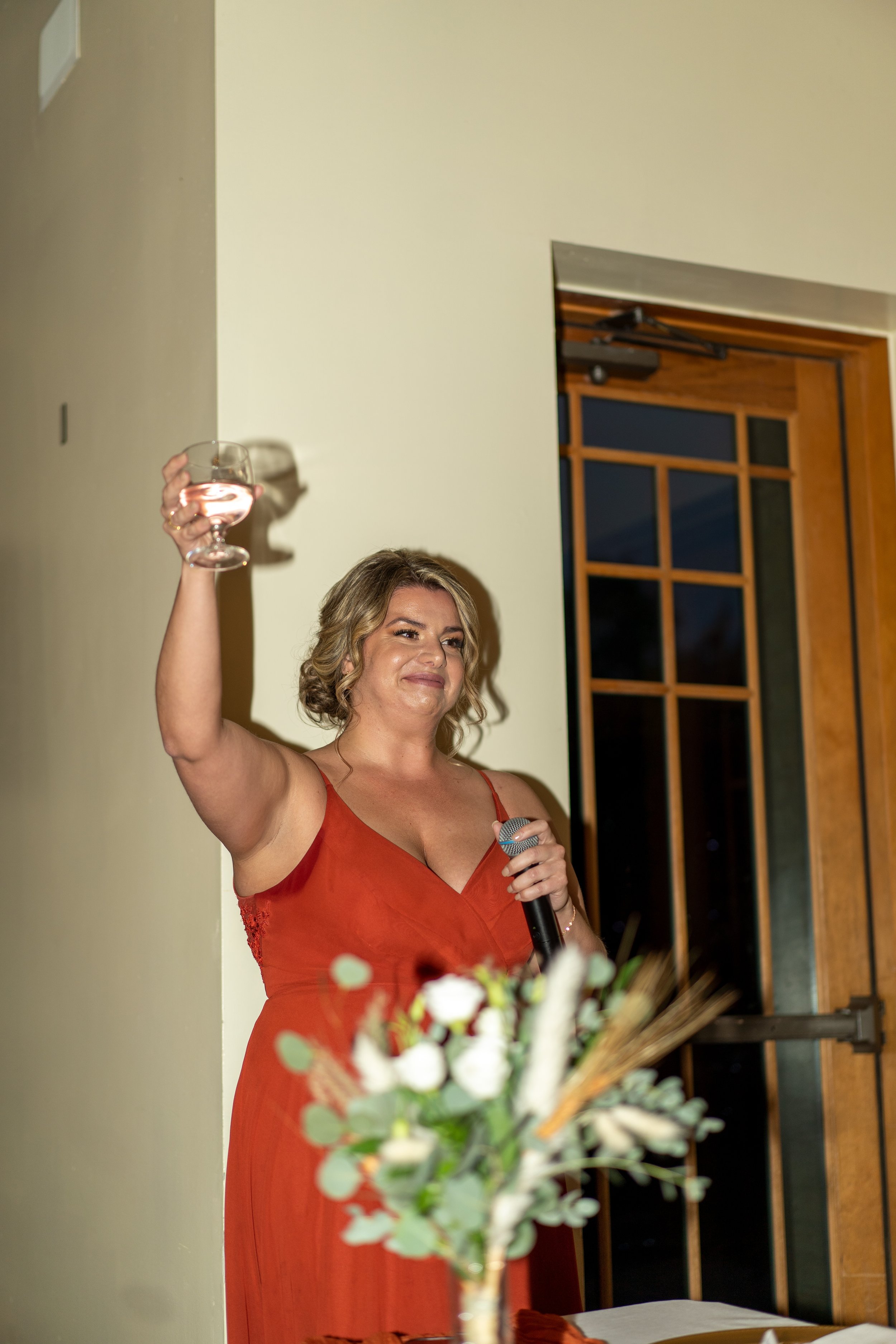 A woman in a red dress holding a glass of wine and a microphone, standing indoors near a doorway with a floral arrangement in front of her.